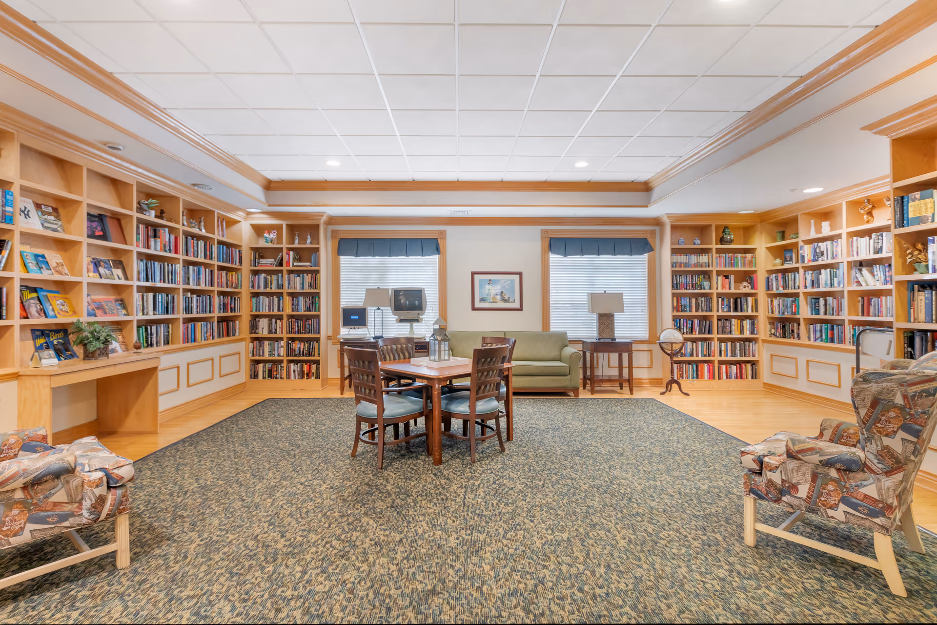 Bright community library room with floor-to-ceiling built-in bookshelves, a central table and chairs, armchairs, and windows.