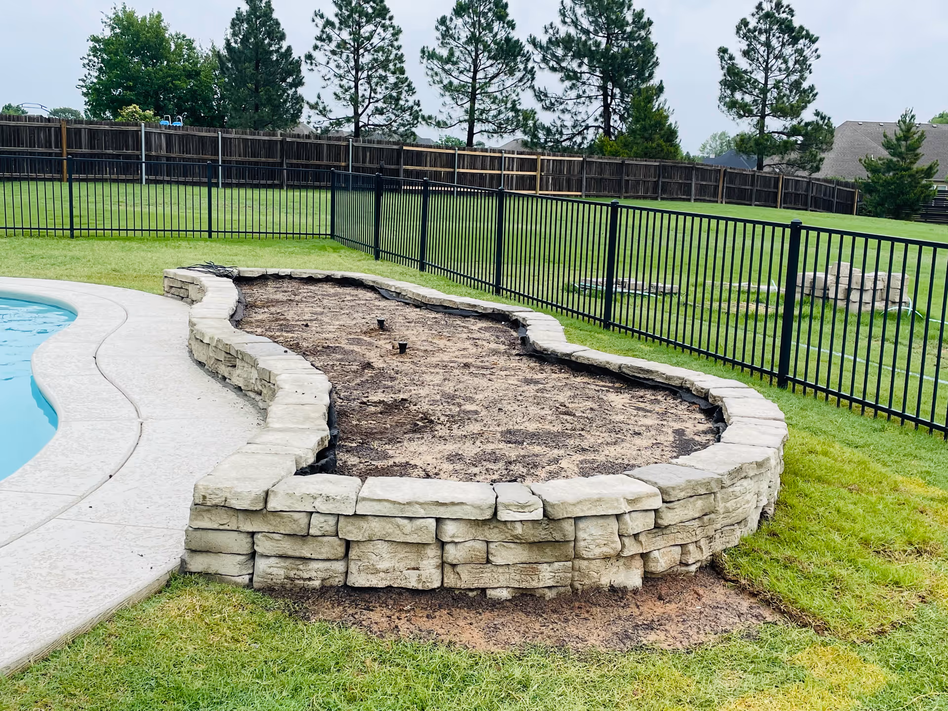 Curved stone planter bed surrounds dirt area beside a pool in a fenced yard.
