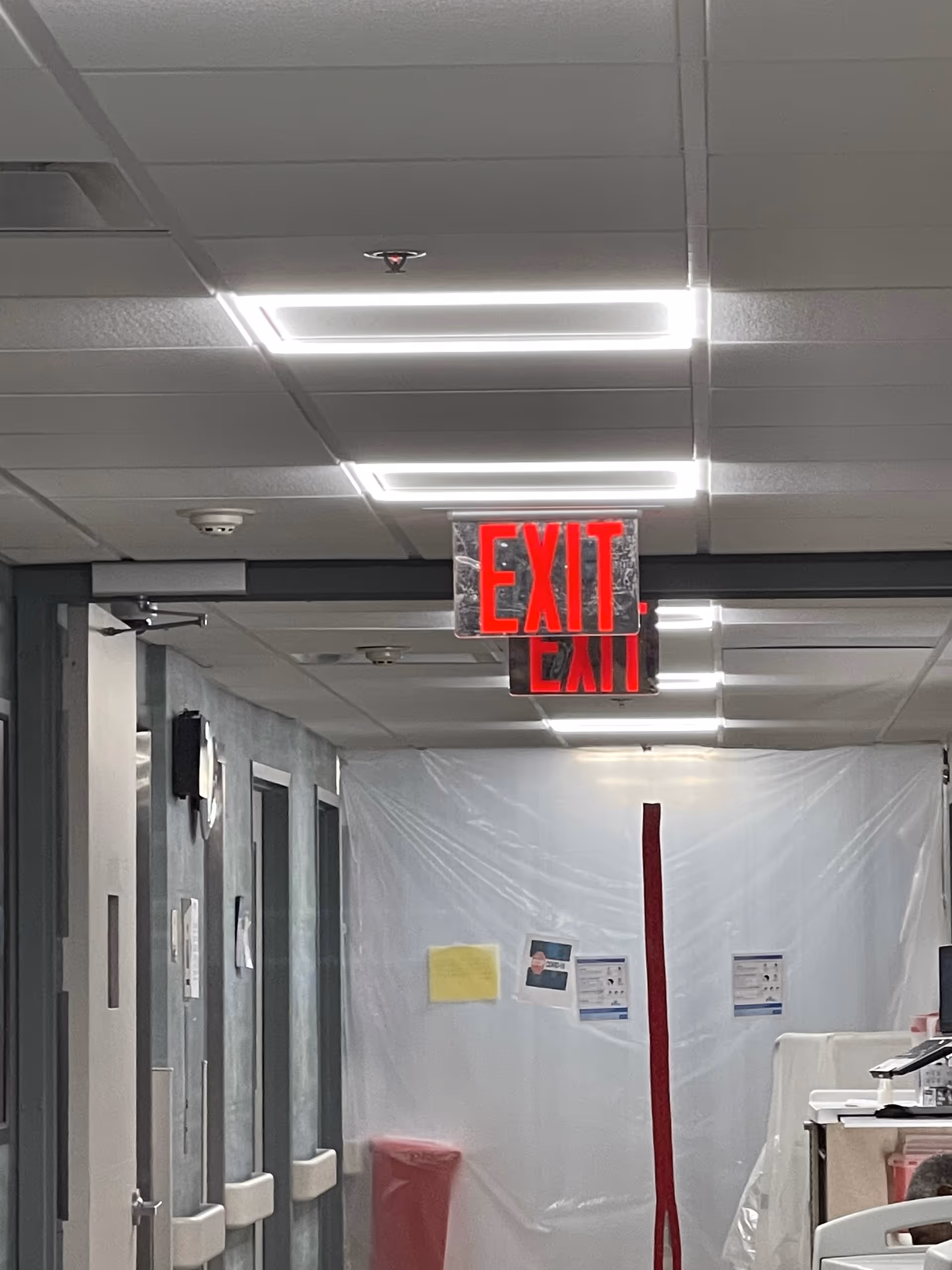 Indoor hospital-like hallway with ceiling lights, exit sign, and plastic barrier.