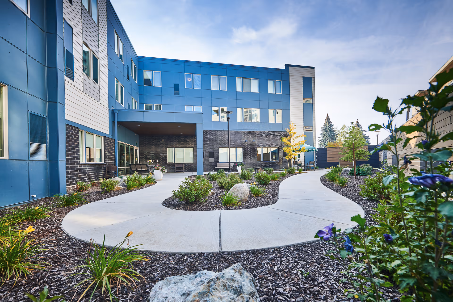 Blue multi-story senior living building with curved walkway and landscaped outdoor courtyard.