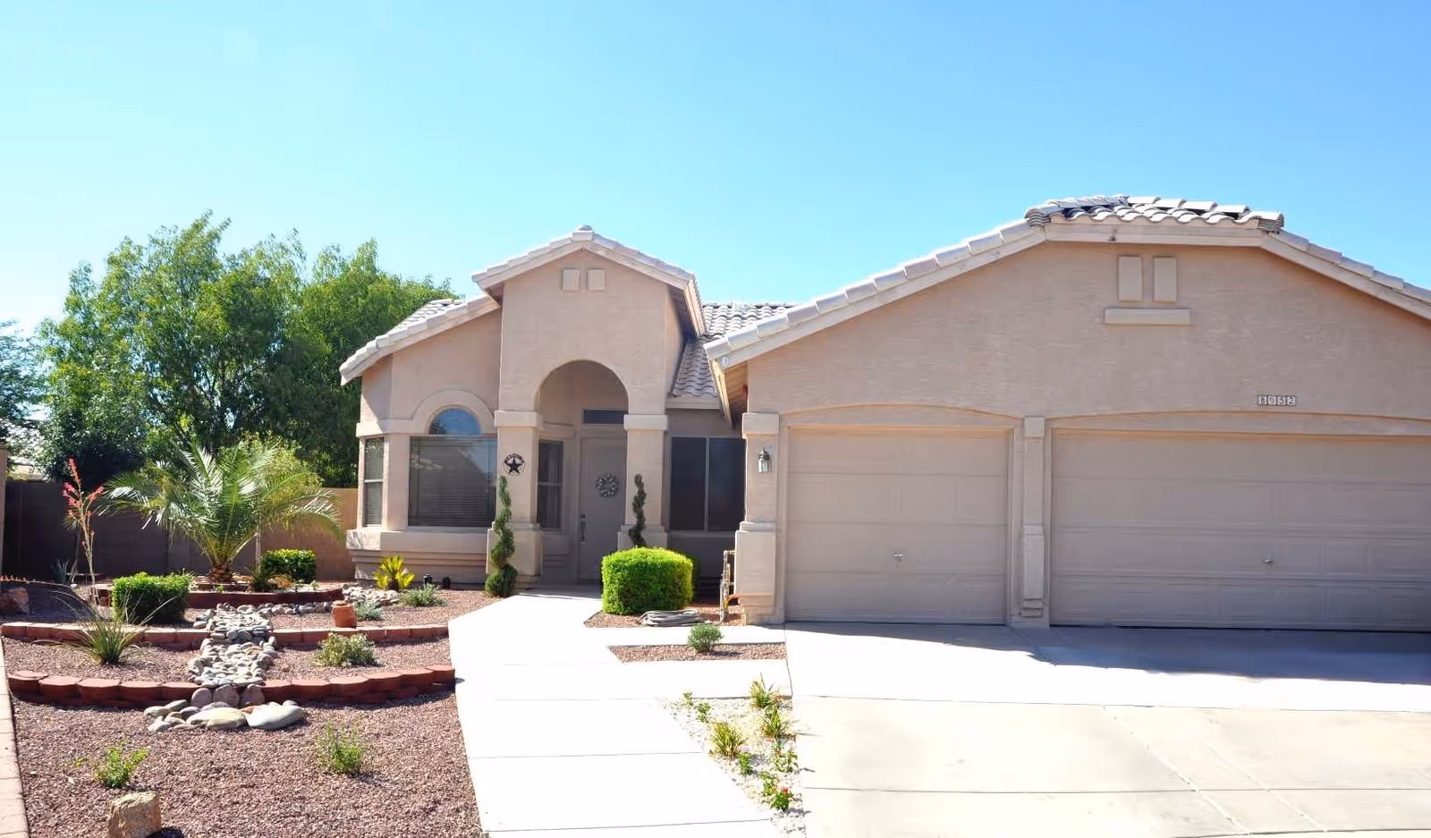 Front exterior of a single-story stucco house with a three-car garage, driveway, walkway and desert landscaping.