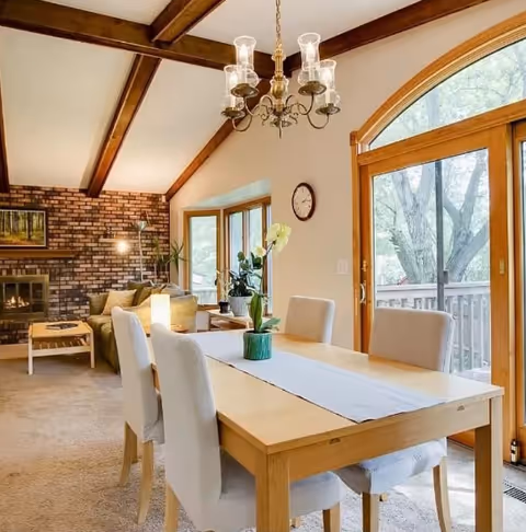 Dining room with wooden table, upholstered chairs, chandelier, windows, and brick fireplace.