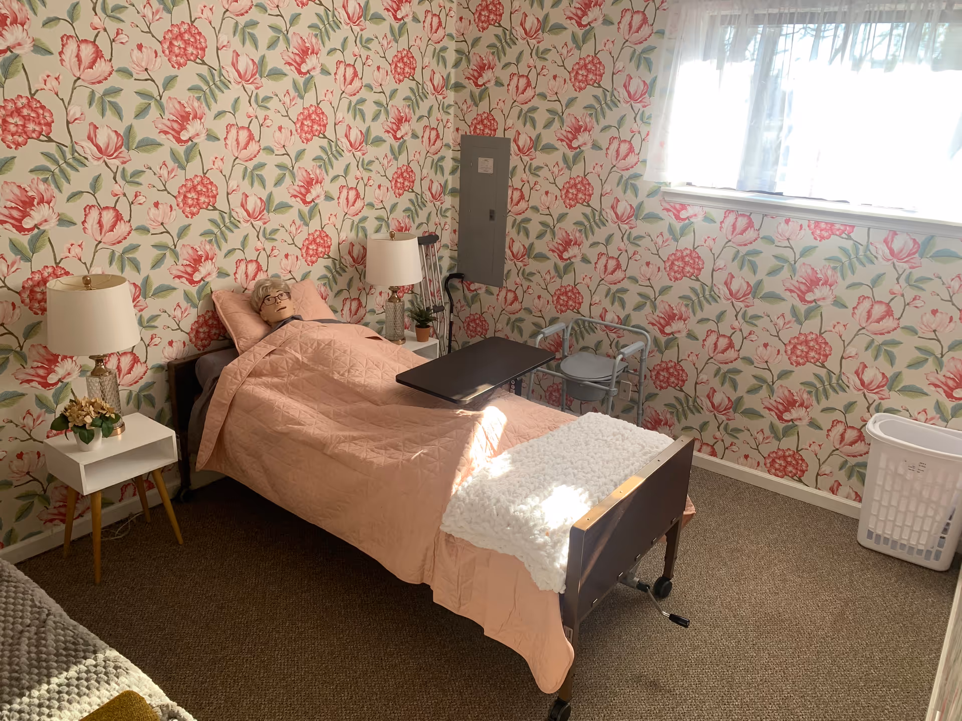 A pink-themed bedroom with a hospital bed, floral wallpaper, lamps, and window light.