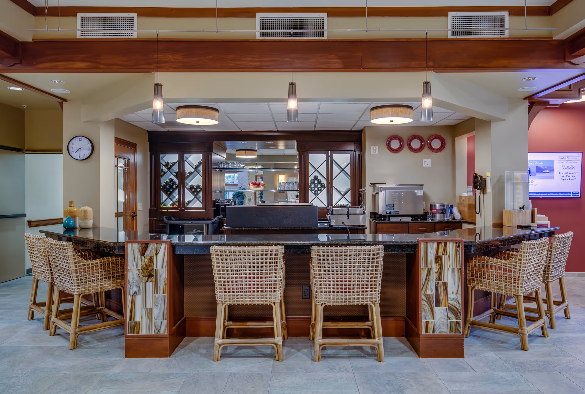 Long granite counter with wicker stools in a senior living dining area, facing glass-front service cabinets.
