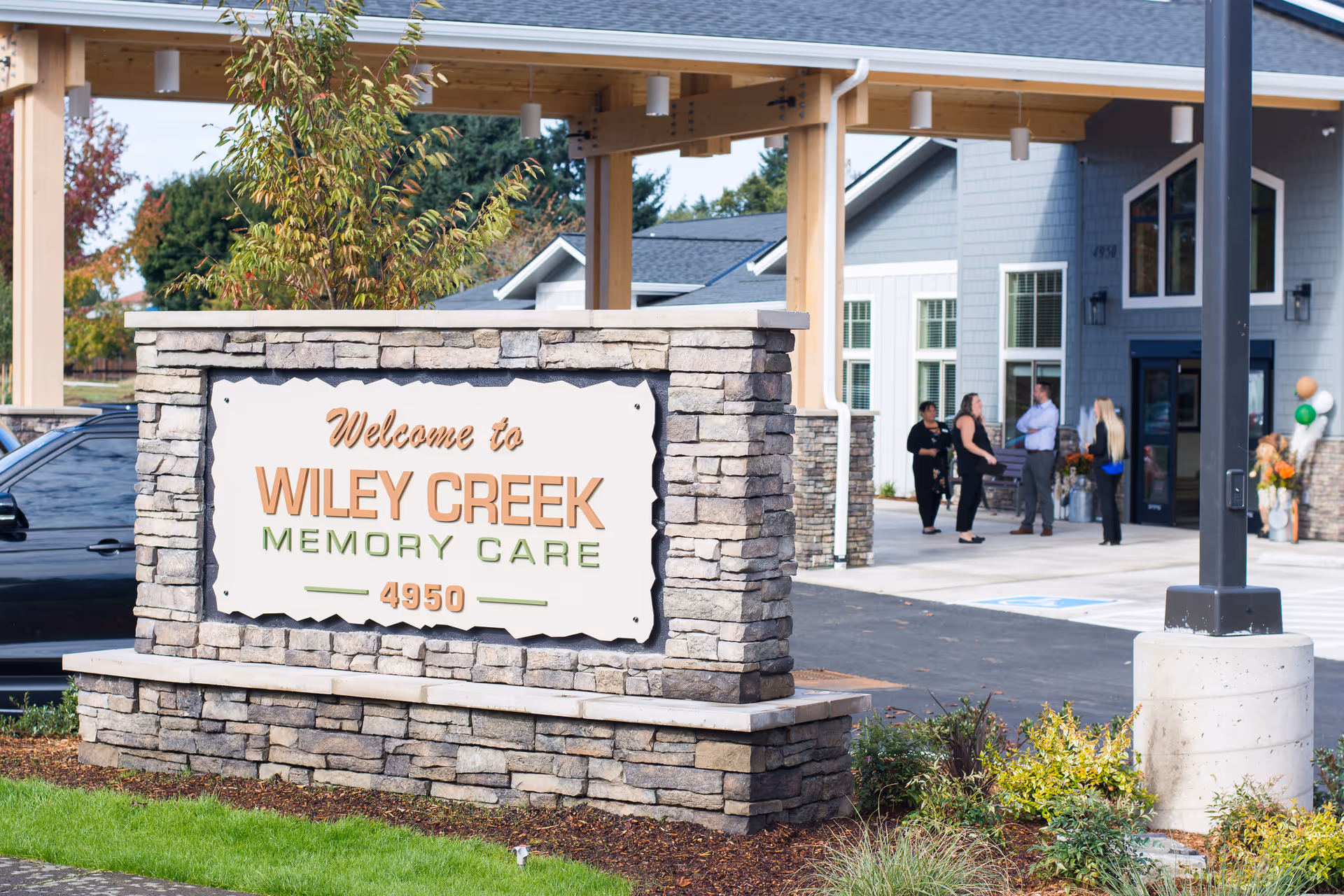Stone sign welcomes visitors to Wiley Creek Memory Care outside the facility entrance with people nearby.