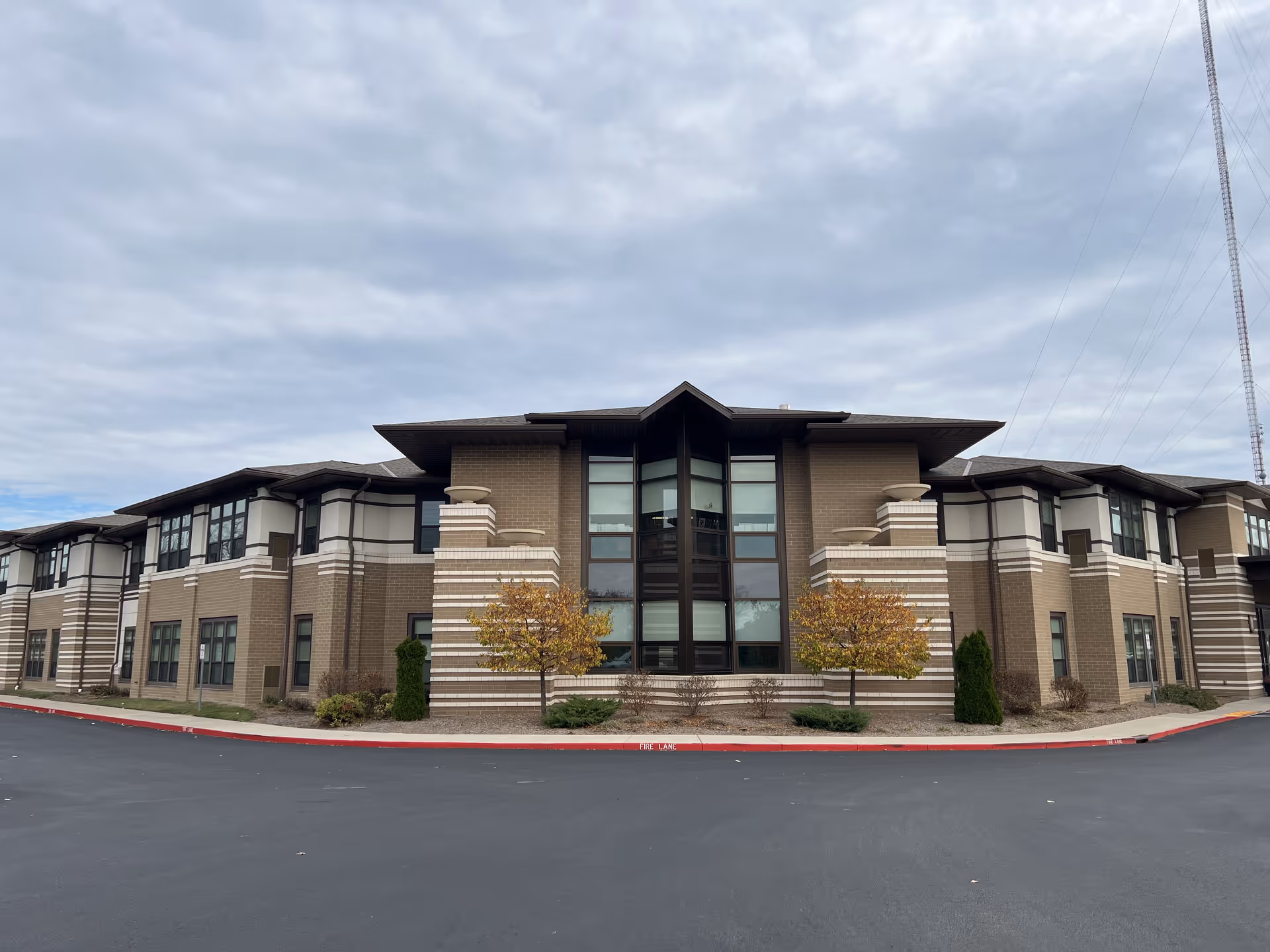 Exterior front view of a two-story senior living facility building with brown and beige brick walls, large windows, and two small trees in front. The sky is cloudy and the pavement in front is freshly paved.