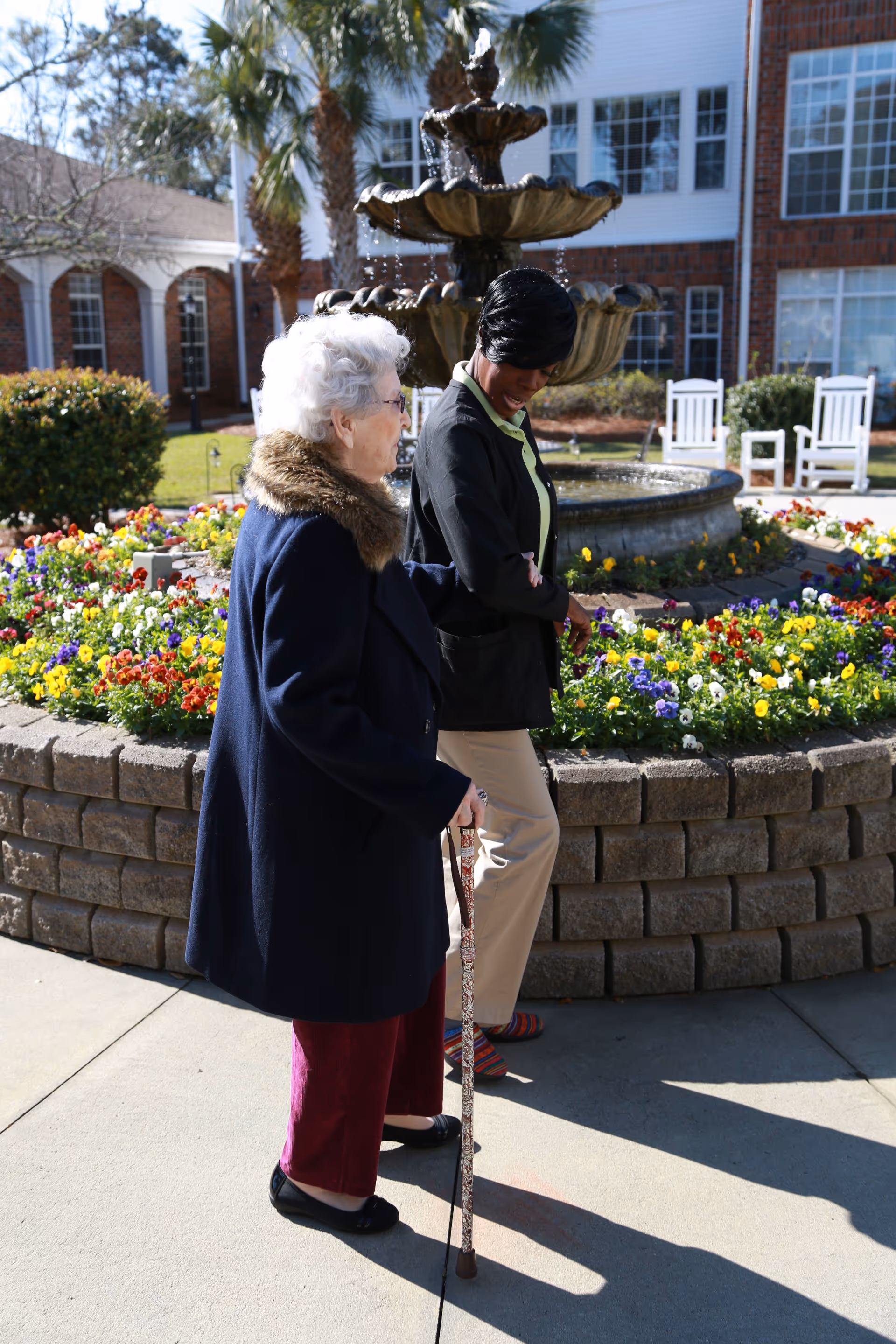 Two women walk by a fountain in a flowered outdoor courtyard at a senior living facility.
