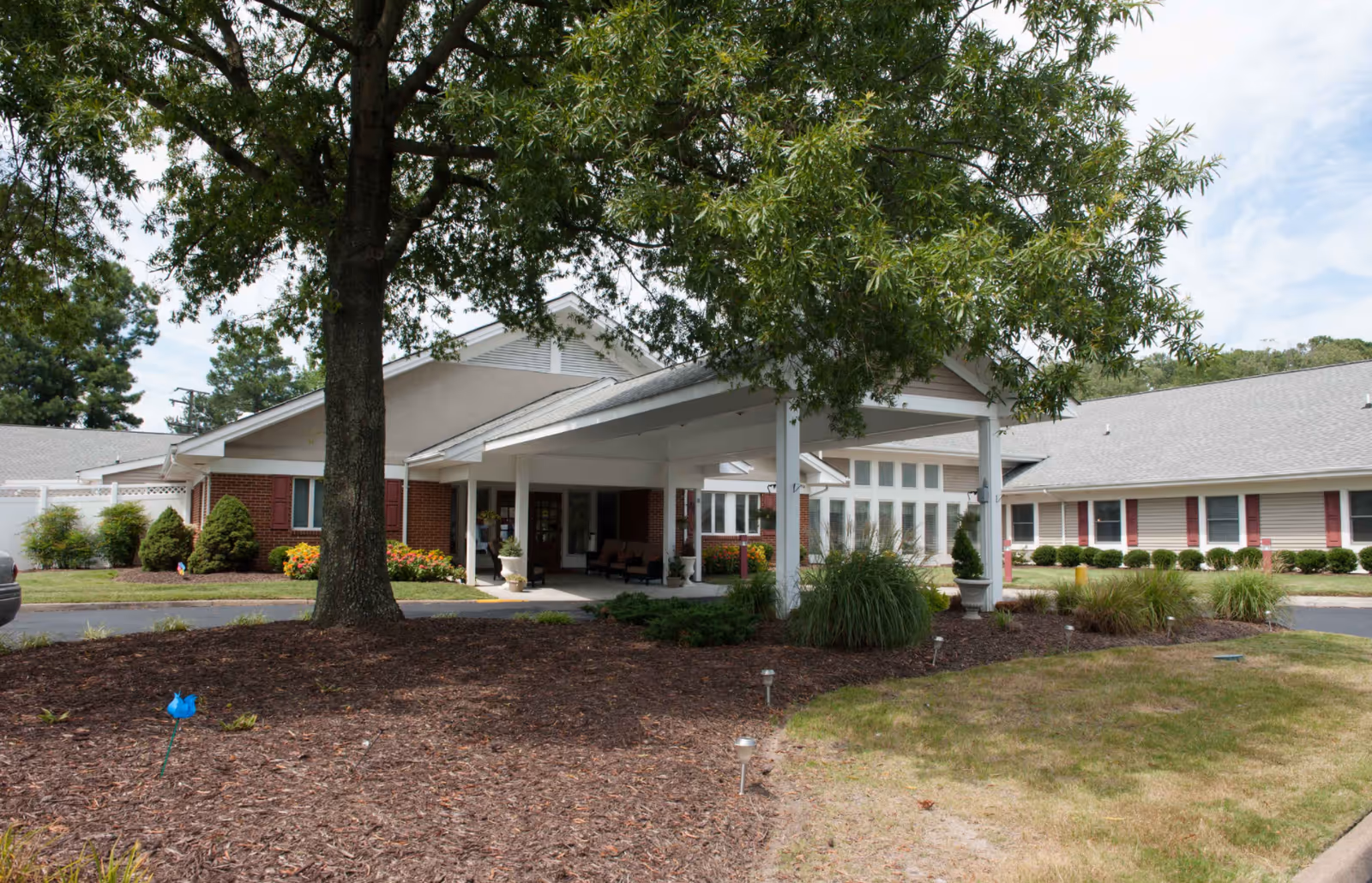 Exterior view of Churchland Nursing Home showing a single-story building with a covered entrance, surrounded by landscaped grounds with trees, bushes, and flower beds under a partly cloudy sky.