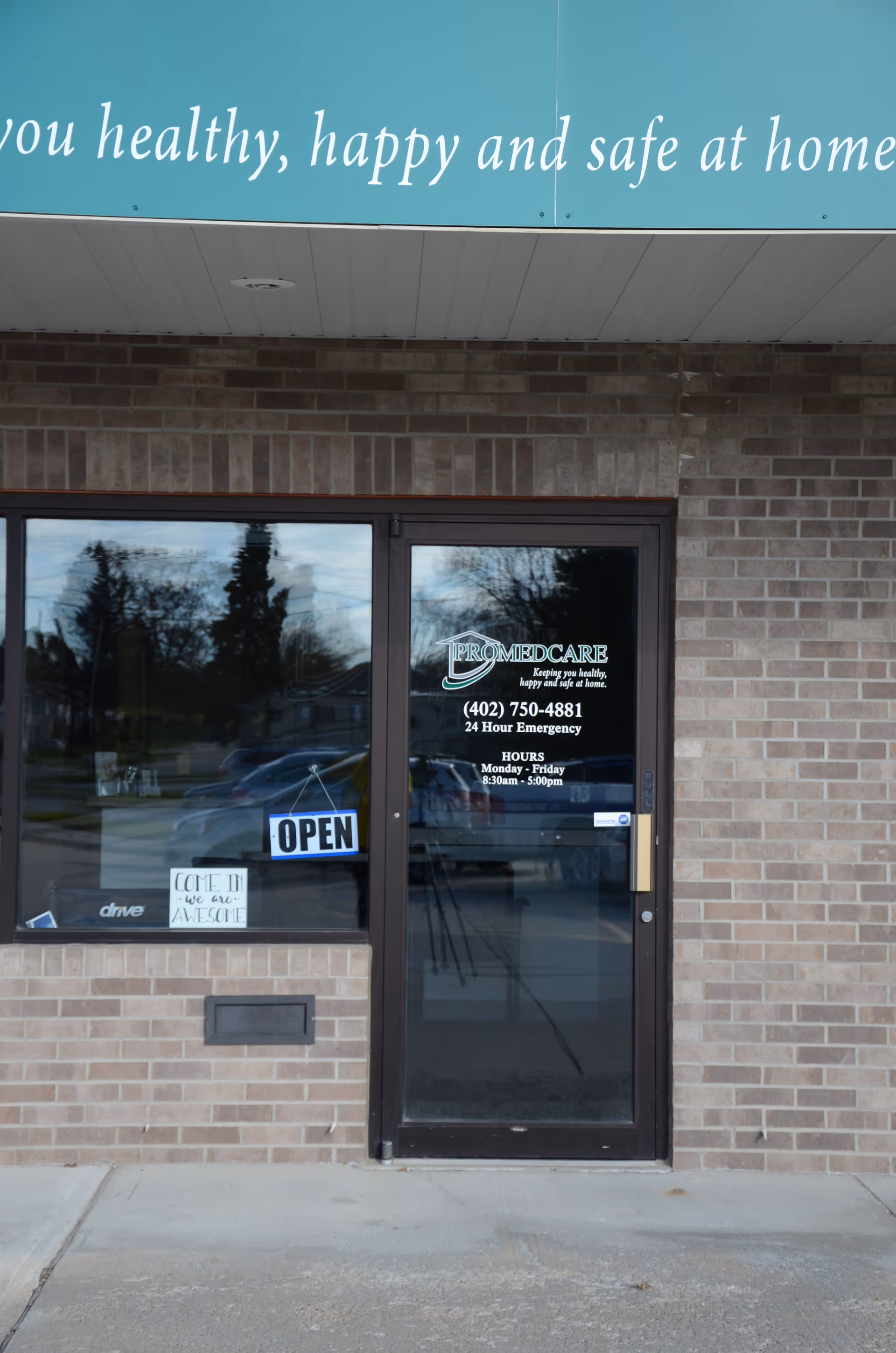 Brick building entrance with a glass door displaying Promedcare branding and an OPEN sign.
