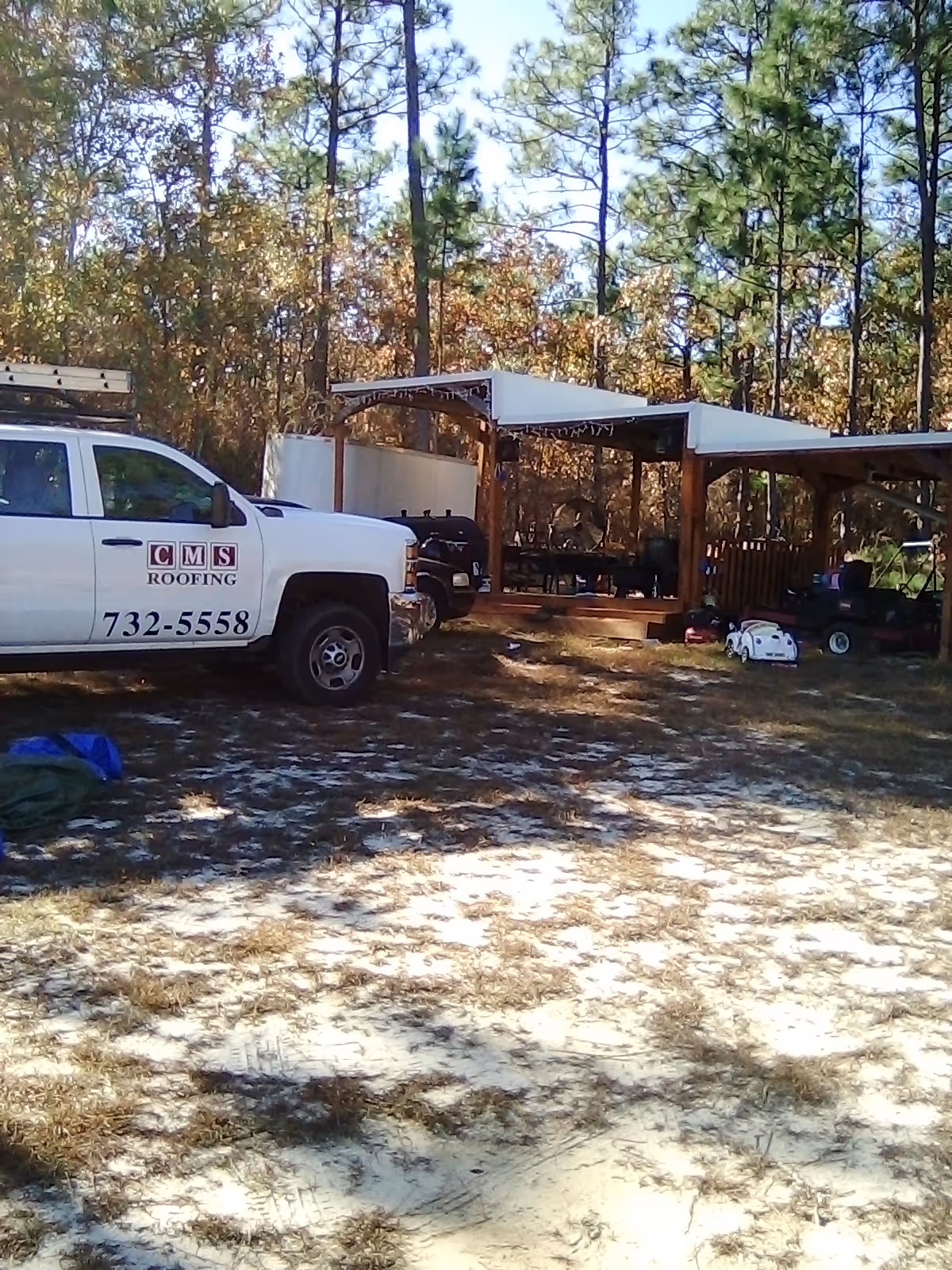 Outdoor site with a CMS Roofing truck and sheltered seating among pines.