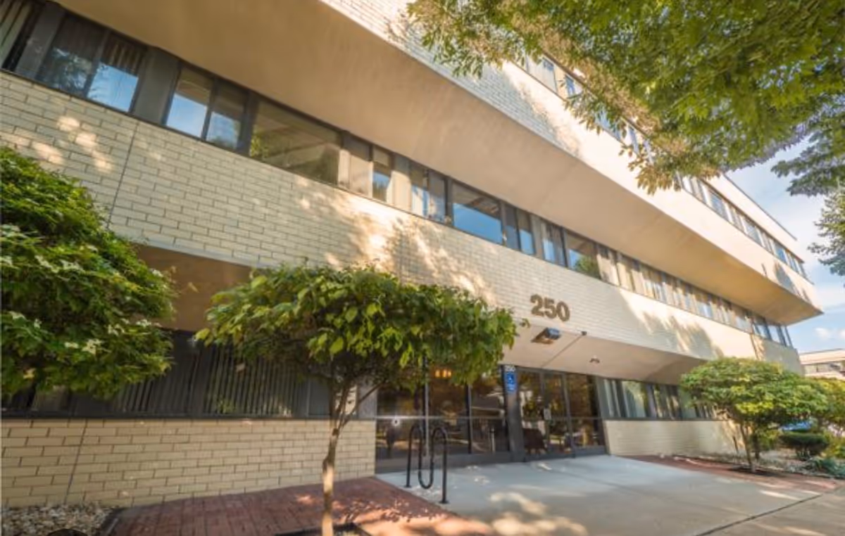Exterior view of a beige brick senior living building with glass entrance and trees.