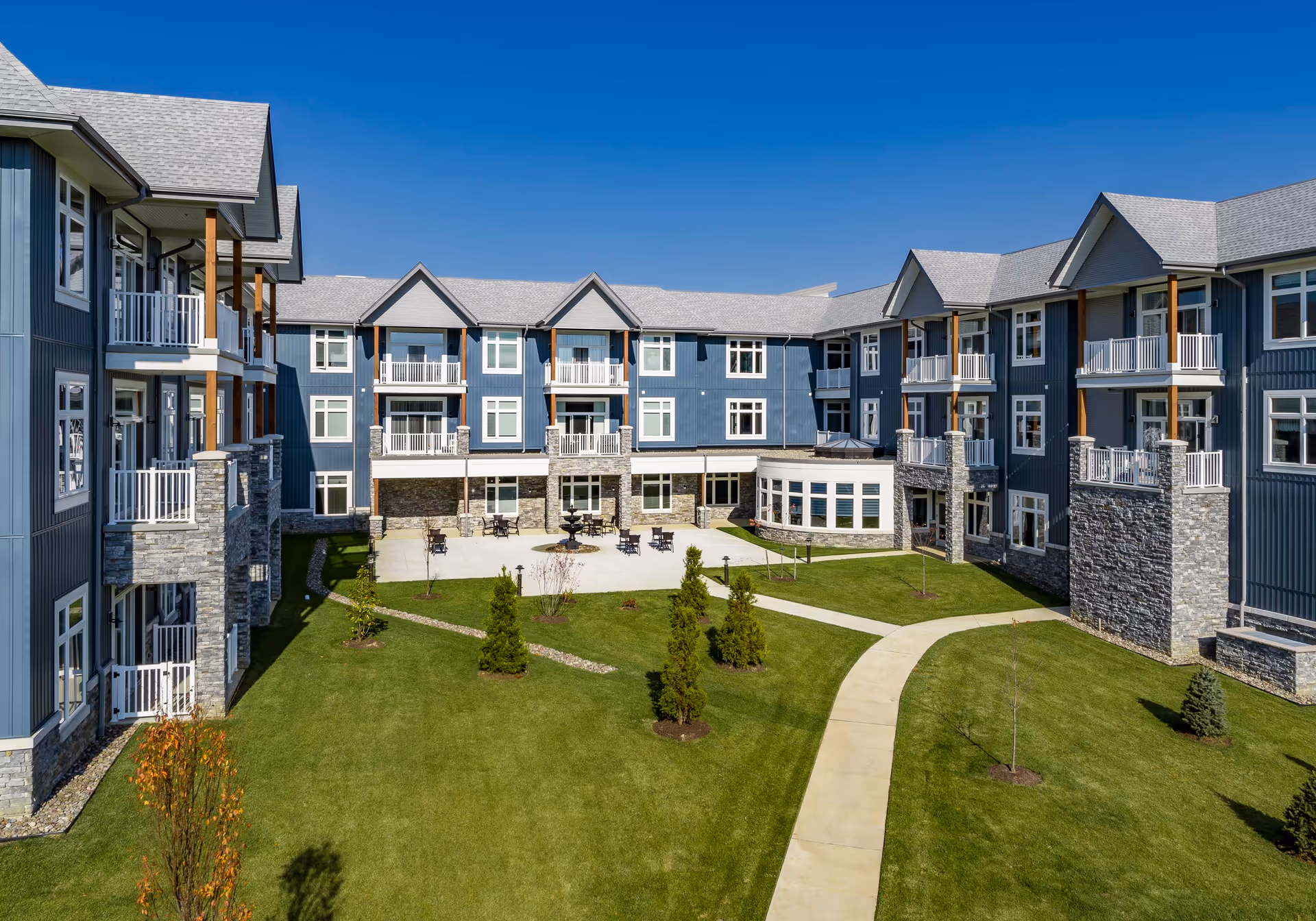 Exterior view of Meadowcrest at Middletown senior living facility showing a large, modern three-story building with blue siding, stone accents, balconies, and a central courtyard with green lawn, small trees, and a paved walkway under a clear blue sky.