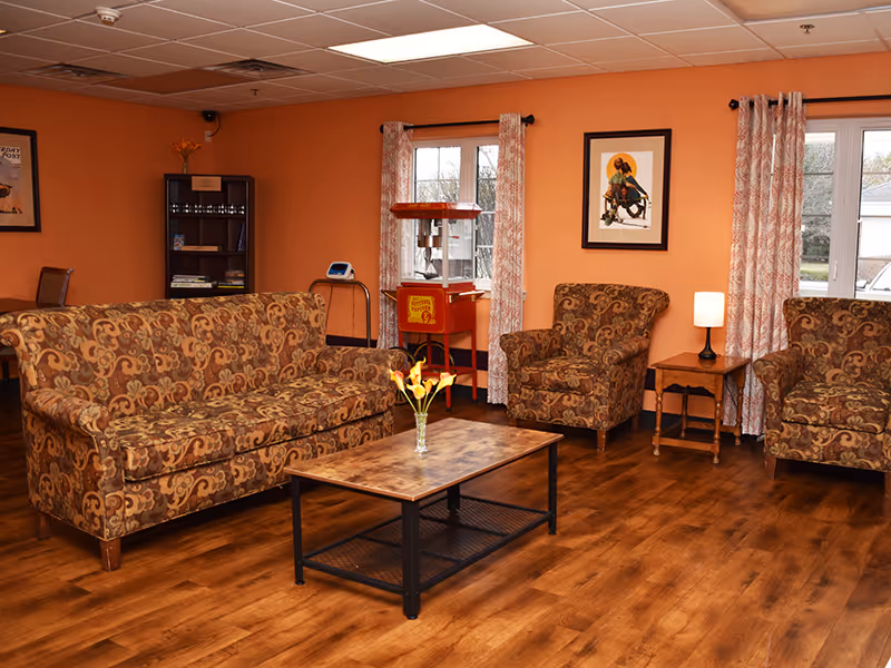 A cozy living room with orange walls, featuring a patterned sofa and two matching armchairs. A wooden coffee table with a vase of yellow flowers is in the center. There is a small side table with a lamp between the armchairs. Two windows with patterned curtains let in natural light. A popcorn machine and a small shelf unit are visible in the background, along with framed artwork on the walls.