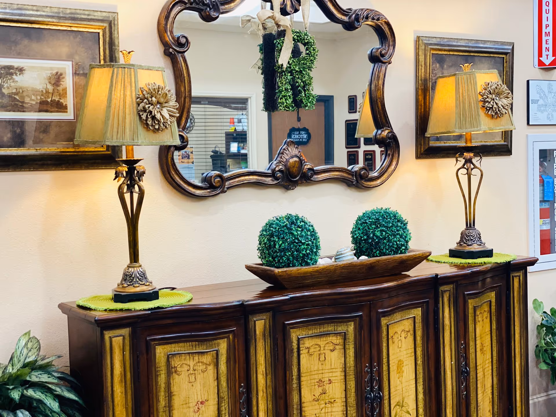 A decorative wooden sideboard with intricate panel designs is topped with two ornate table lamps with beige shades, a wooden tray holding two green spherical topiary plants, and a large ornate mirror hanging on the wall above. The wall also features framed artwork and a fire equipment sign, with some indoor plants visible at the edges.