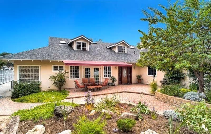 Front exterior of a single-story house with a paved patio, outdoor dining set, and landscaped garden.