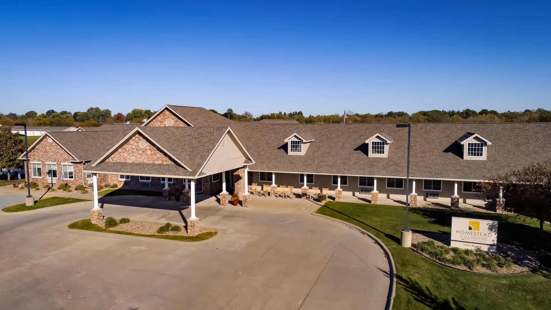 Exterior view of Homestead Assisted Living & Memory Care of Oskaloosa, showing a single-story building with a covered entrance, stone and siding facade, a driveway, and a sign with the facility name on a well-maintained lawn under a clear blue sky.