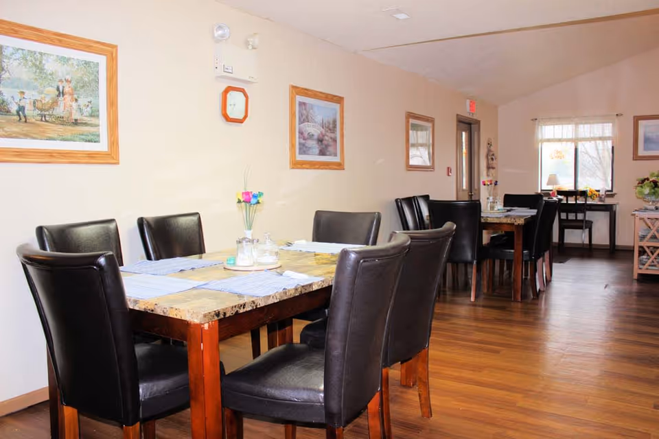 Interior view of a dining area with two wooden tables, each surrounded by black leather chairs. The tables have placemats, a small vase with colorful artificial flowers, and condiments. The room has wooden flooring, light-colored walls adorned with framed paintings, and a window with sheer curtains letting in natural light.