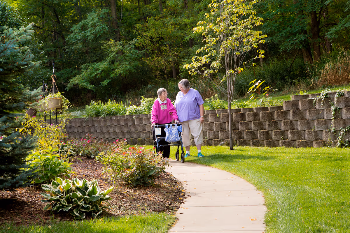 Two elderly women walking along a curved garden path surrounded by green grass, plants, and trees. One woman is using a walker and wearing a pink jacket, while the other woman, dressed in a light purple top and beige pants, walks beside her. A stone retaining wall and lush greenery are visible in the background.