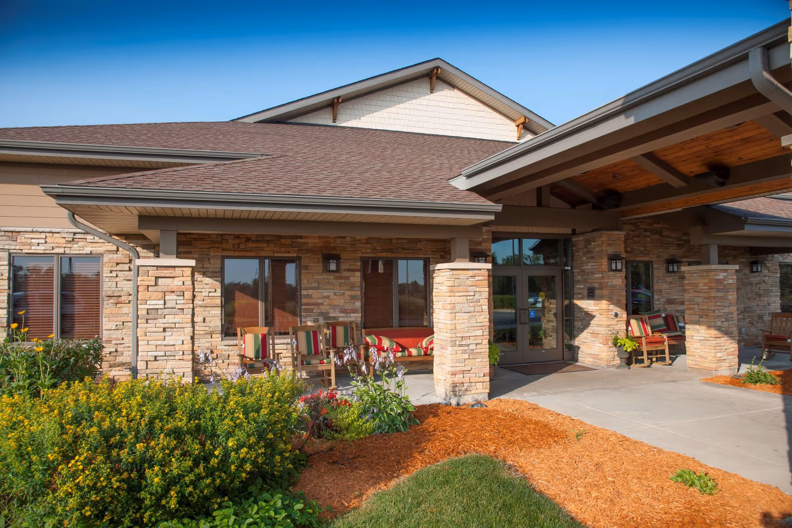 Exterior view of NorthPointe Terrace facility showing a covered entrance with stone pillars, outdoor seating with striped cushions, and landscaped garden beds with green shrubs and flowers under a clear blue sky.