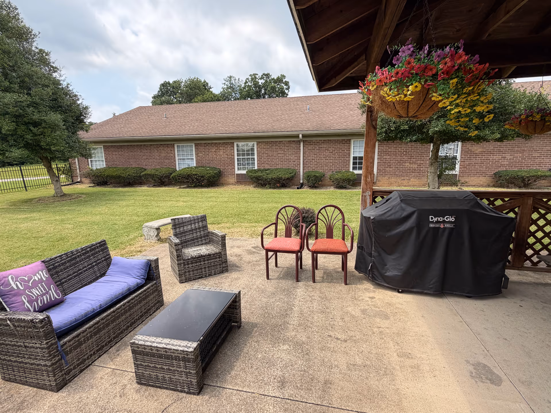 Outdoor patio with wicker seating, red chairs, hanging flowers, covered grill beside brick building.