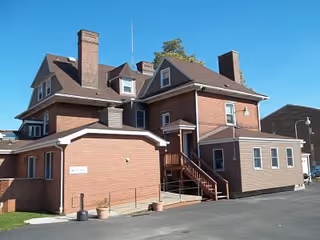 Exterior view of a large multi-story brick building with multiple chimneys and a small staircase leading to an entrance. The sky is clear and blue.