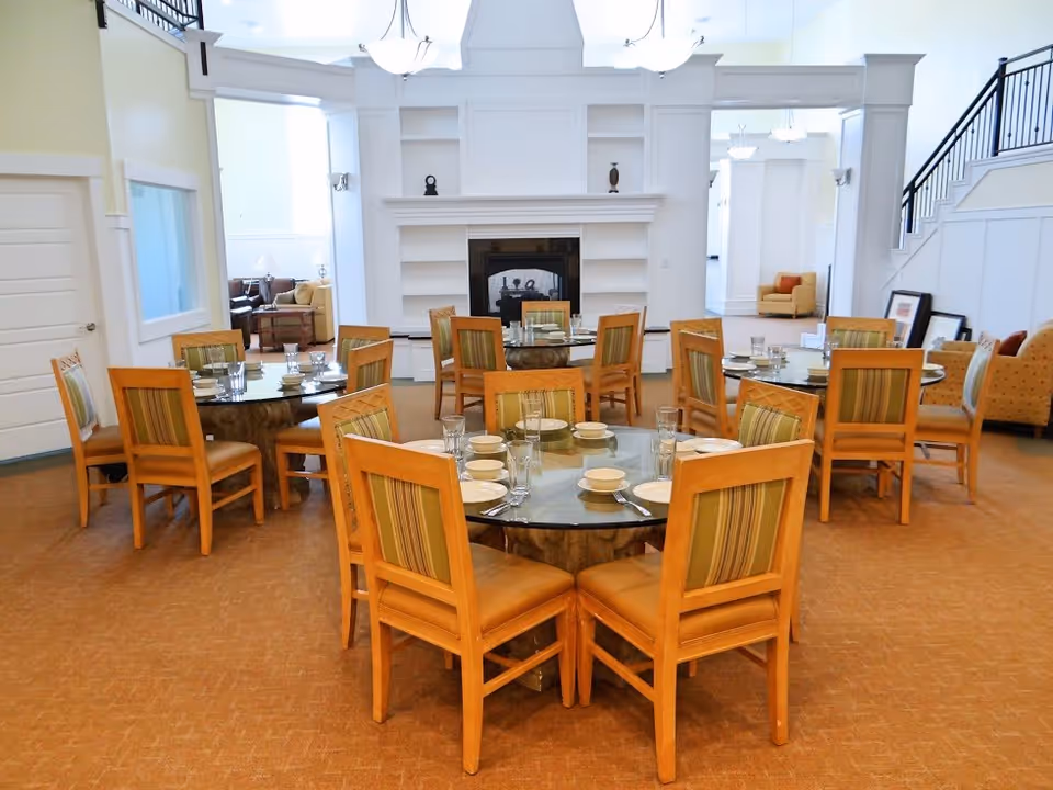 A bright dining room with multiple round tables set with plates, bowls, glasses, and silverware. Each table is surrounded by wooden chairs with striped cushions. The room features a fireplace with built-in shelves and a high ceiling with hanging light fixtures. There are seating areas with armchairs visible in the background near a staircase.