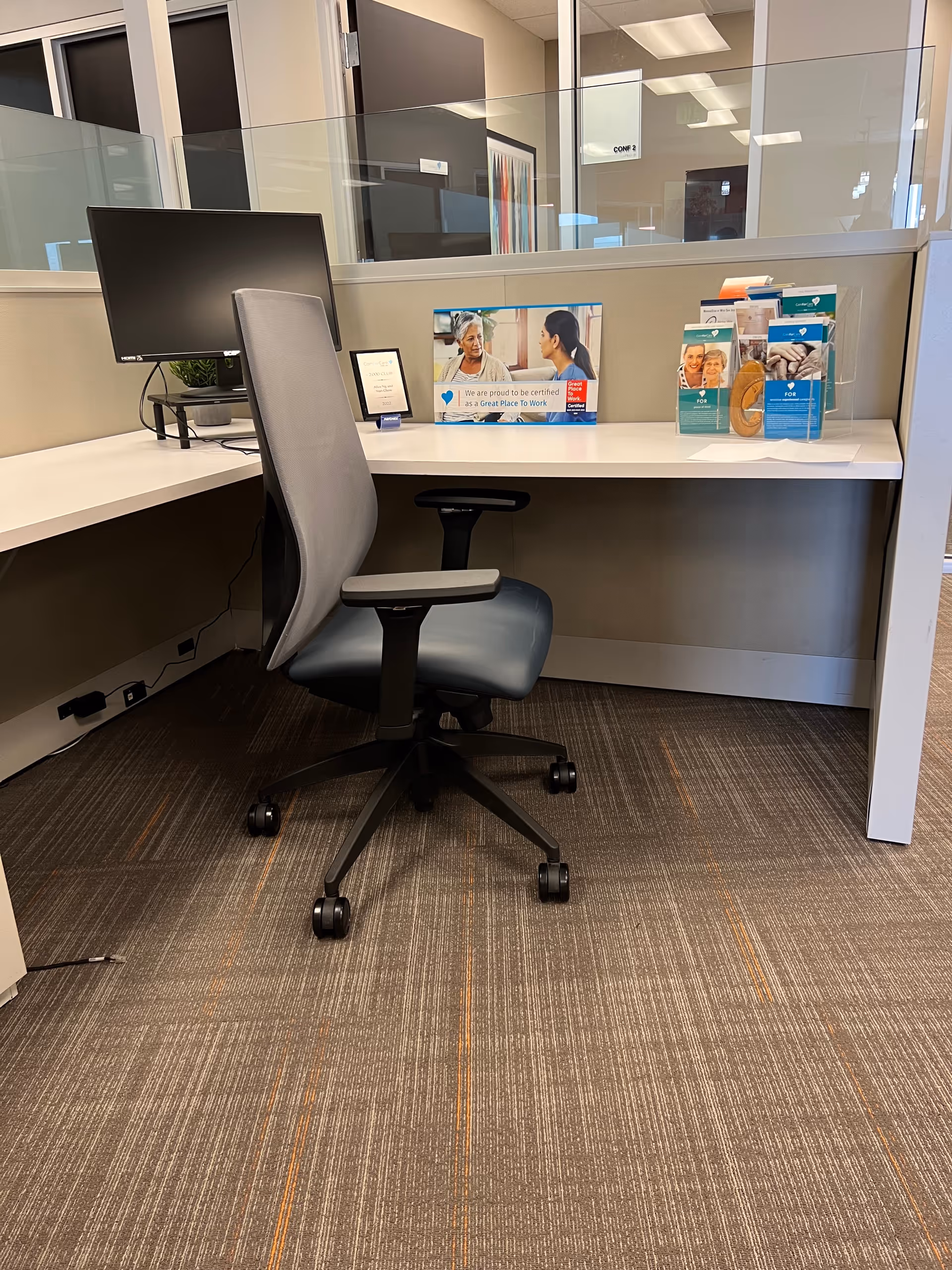 Office cubicle with monitor, chair, and brochures on a white desk near glass partition.