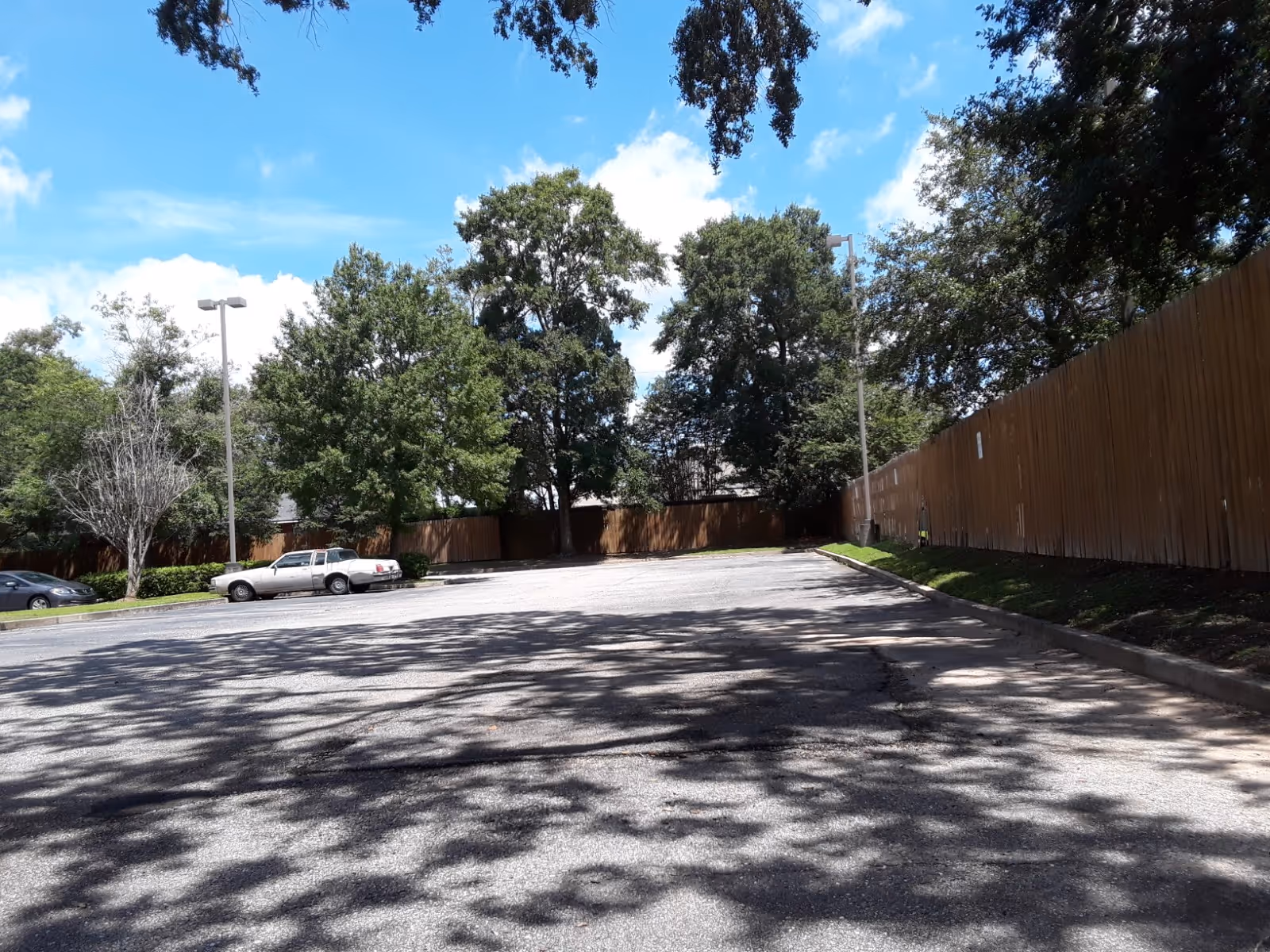 Parking lot bordered by tall wooden fence, trees, and a few parked cars.
