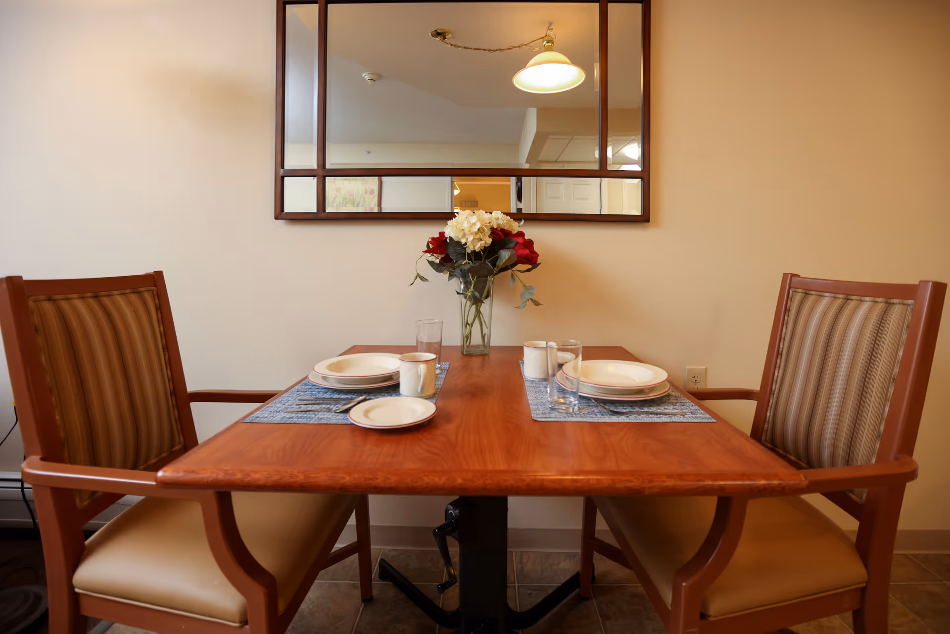 A small dining table set for two with plates, cups, glasses, and a vase of flowers in the center. Two wooden chairs with cushioned seats and striped backs are positioned on either side of the table. A large rectangular mirror hangs on the wall behind the table, reflecting a ceiling light and part of the room.