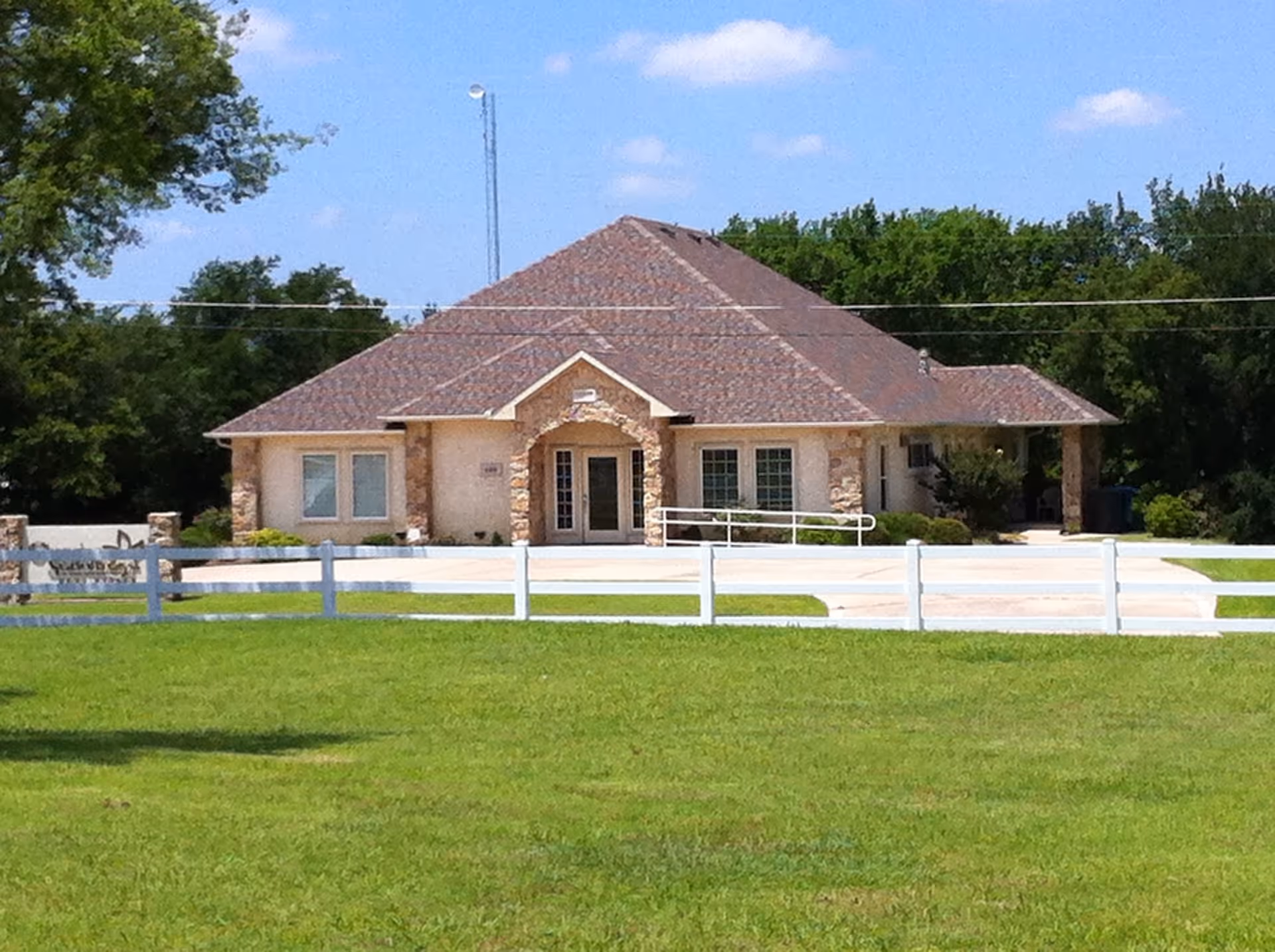A single-story beige building with stone columns, curved entrance, and white fence on green lawn.