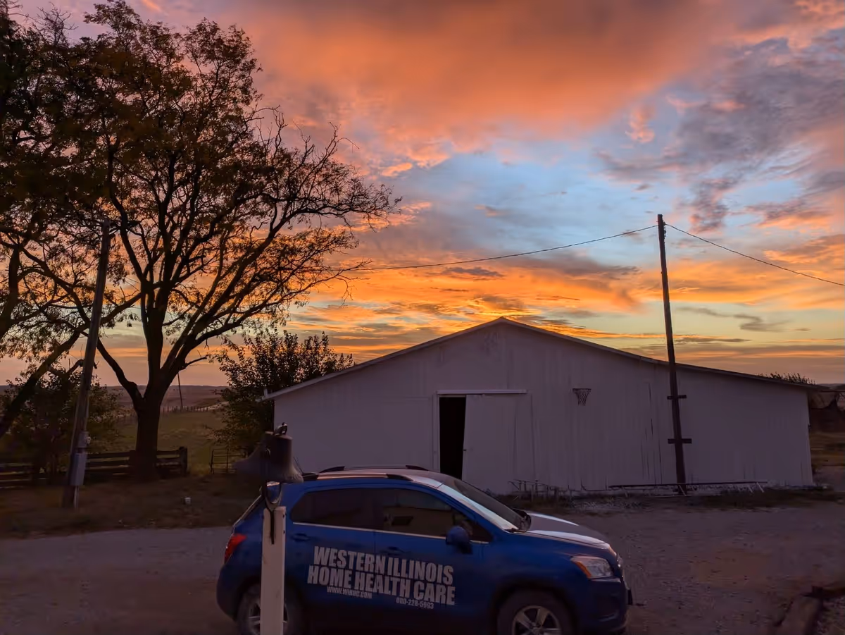 Sunset sky over a white barn with a blue car labeled Western Illinois Home Health Care.