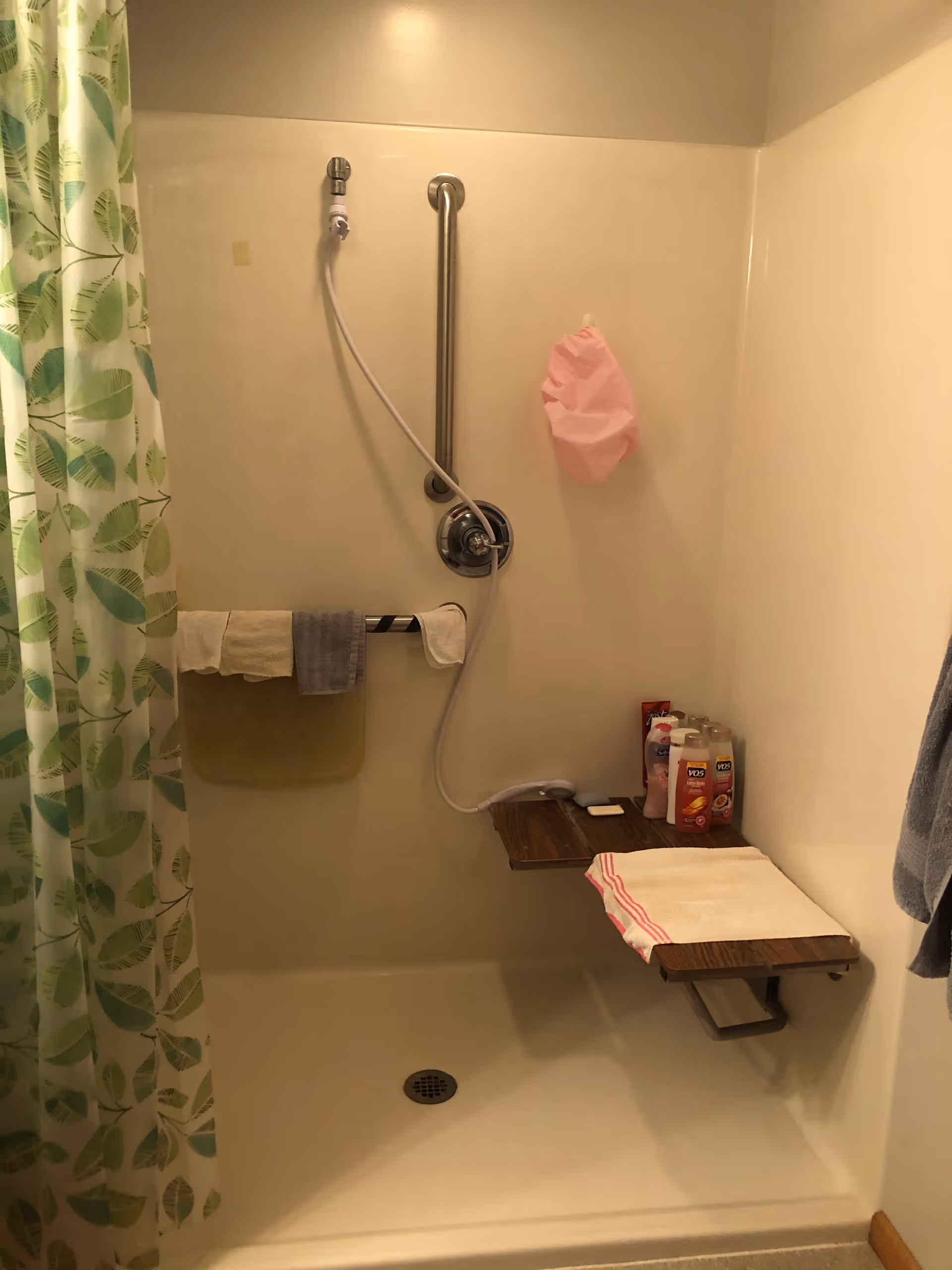 A shower area with a green leaf-patterned shower curtain on the left. Inside the shower, there is a handheld showerhead attached to a vertical grab bar. A small wooden fold-down seat is mounted on the right wall, with a white towel with red stripes placed on it. Several bottles of shampoo and body wash are on the seat. A towel rack with three towels is mounted on the back wall, and a pink shower cap or bag hangs on the right wall.