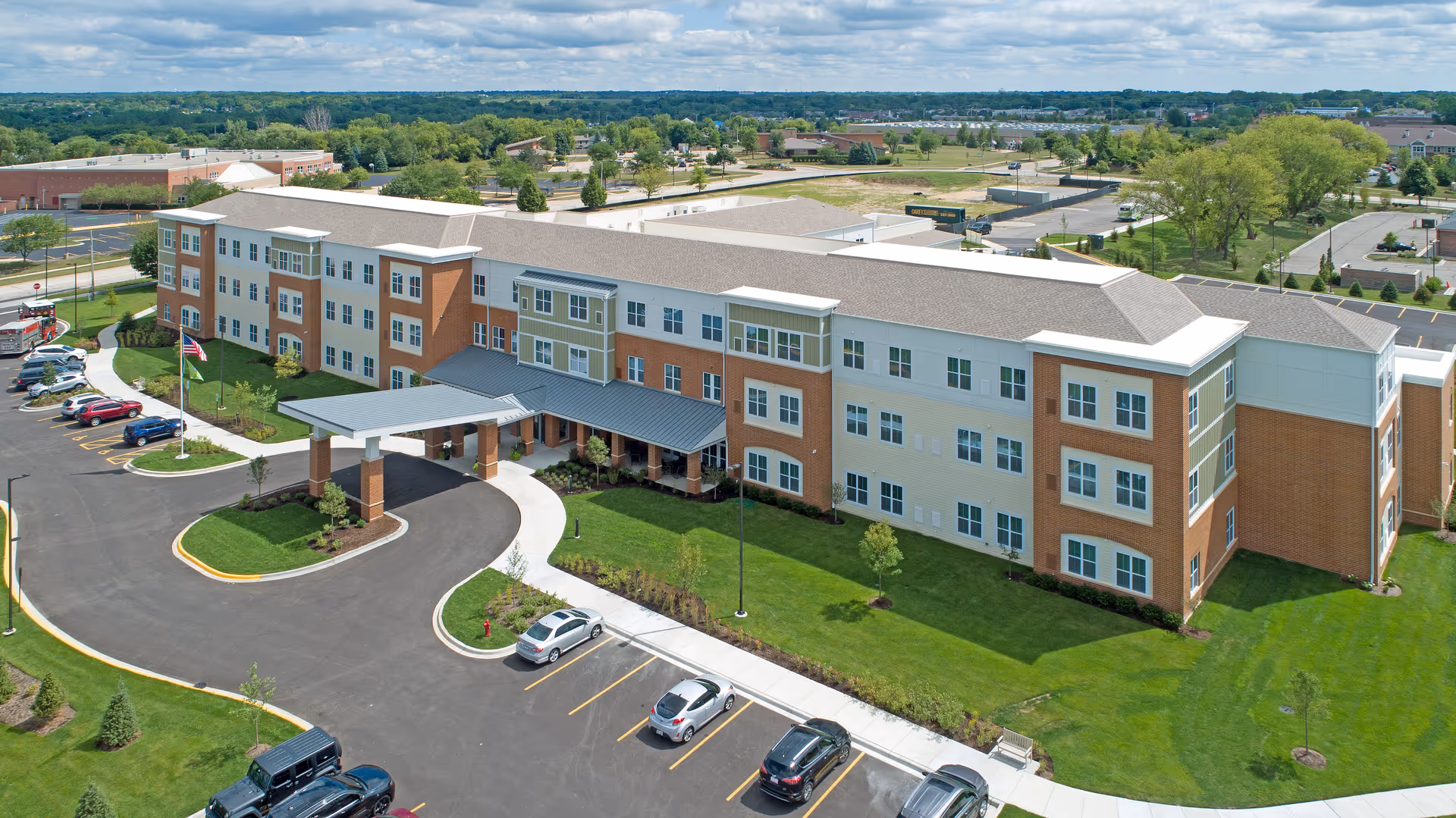 Aerial view of a three-story assisted living facility with a covered entrance, parking lot, and landscaped grounds.