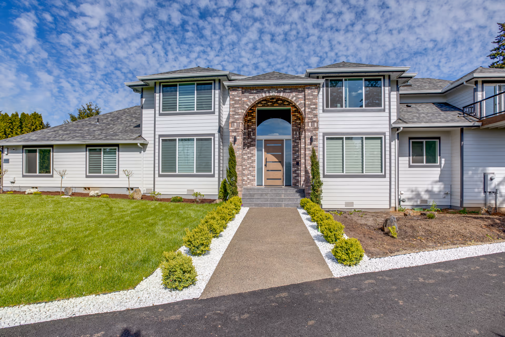 Front exterior of a modern two-story senior living building with a central arched entrance, walkway, and landscaped lawn under a blue sky.