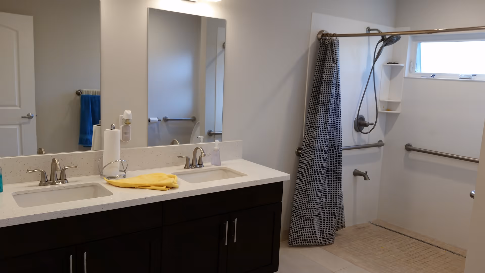 A bathroom with a double sink vanity featuring two faucets, a large mirror above, and a yellow cloth on the counter. To the right is a walk-in shower with a handheld showerhead, grab bars, a small window, and a black and white patterned shower curtain.