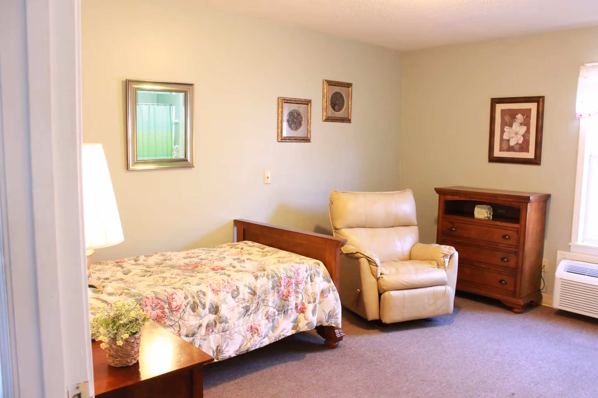 A cozy senior living bedroom with a floral bedspread, recliner, and wooden dresser.
