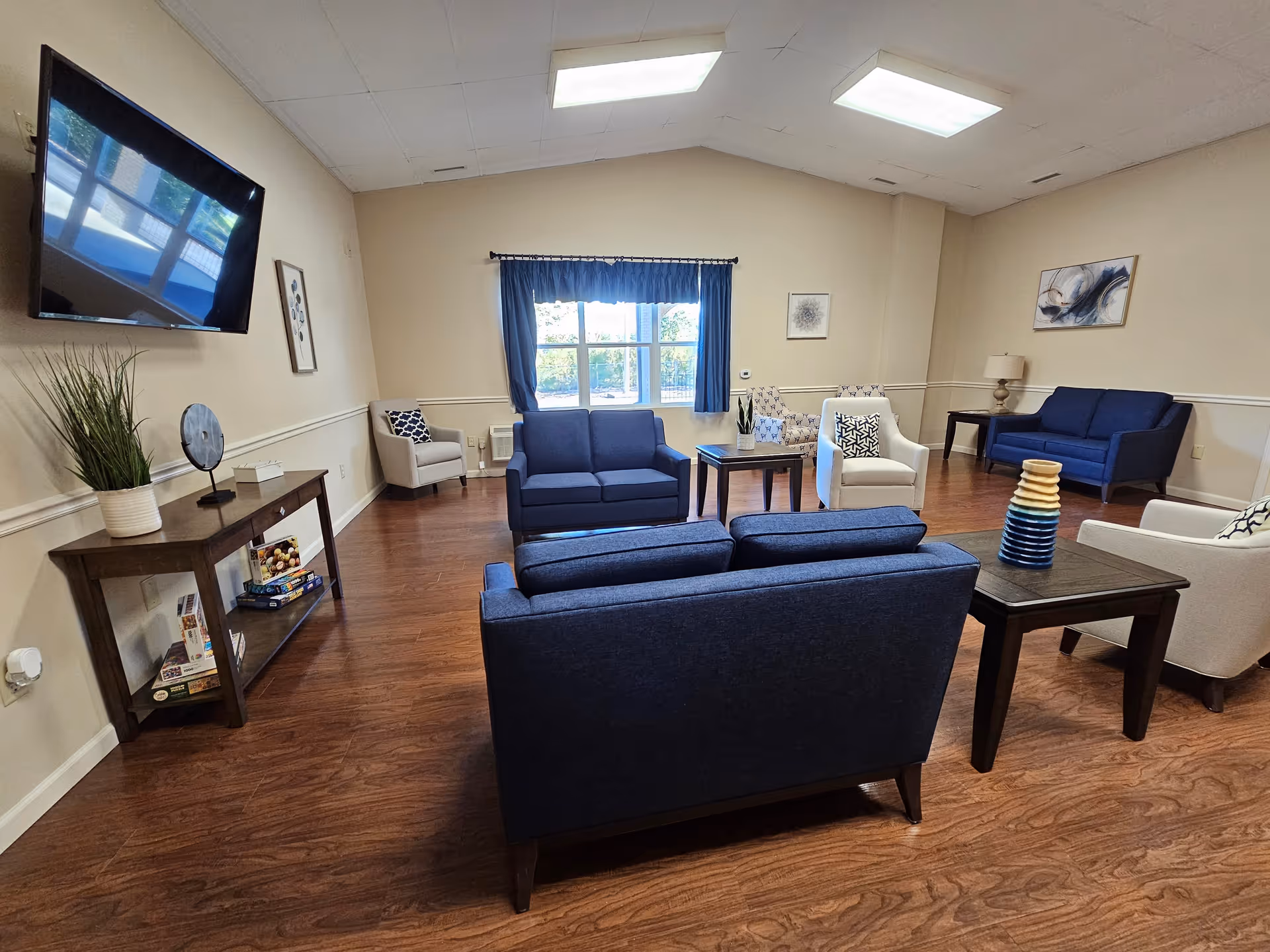 A spacious living room with wooden flooring, featuring multiple blue and beige armchairs and sofas arranged around dark wooden coffee tables. A large window with blue curtains lets in natural light. A flat-screen TV is mounted on the wall above a console table with decorative items and board games. The walls are beige with simple artwork and a table lamp on a side table.