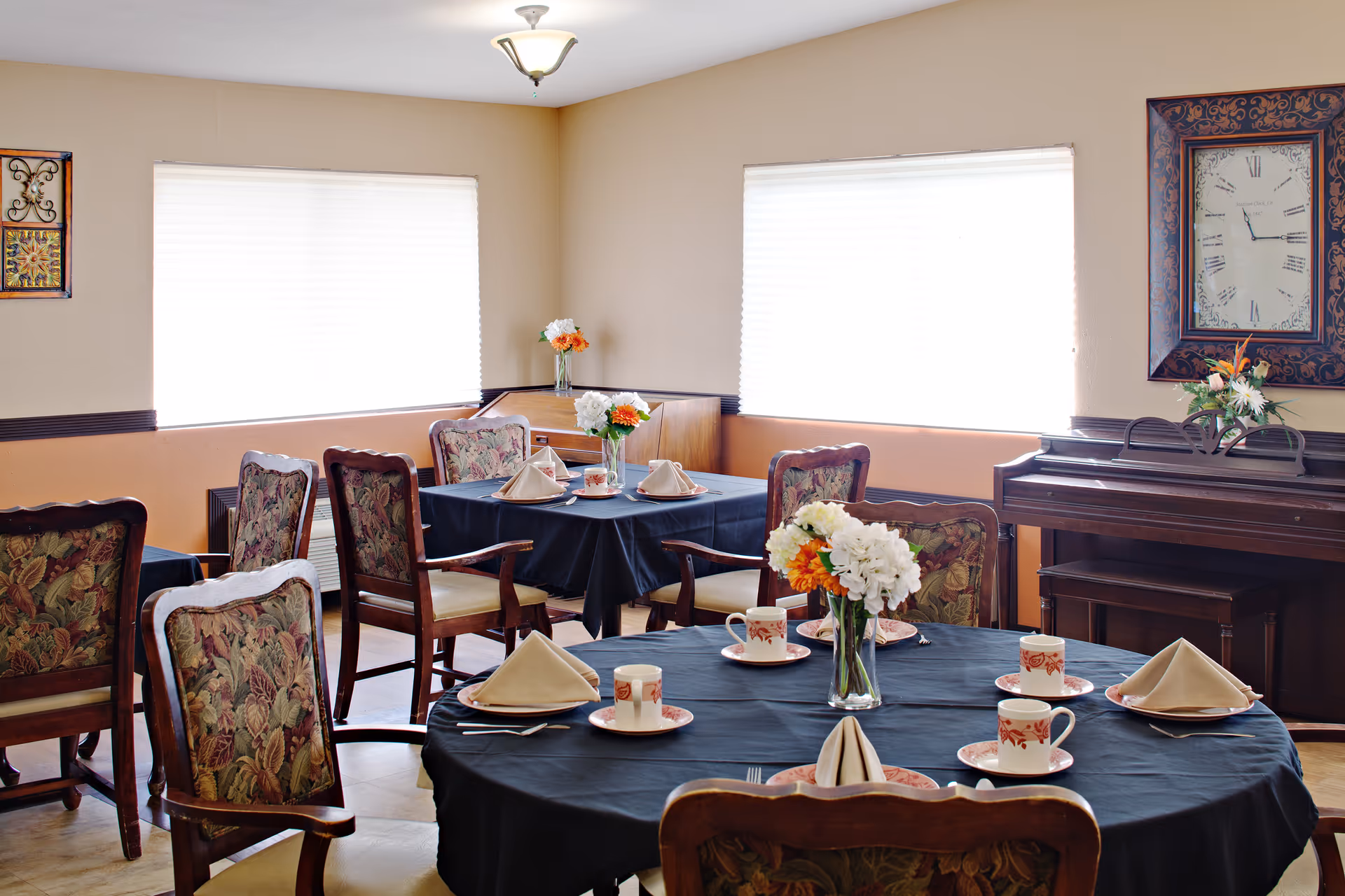 Bright dining room with round tables, floral chairs, and a piano along the wall.