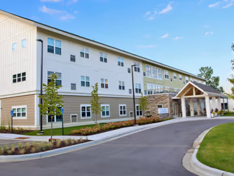 Front exterior of a three-story senior living building with a covered entrance, driveway, and landscaped grounds under a blue sky.