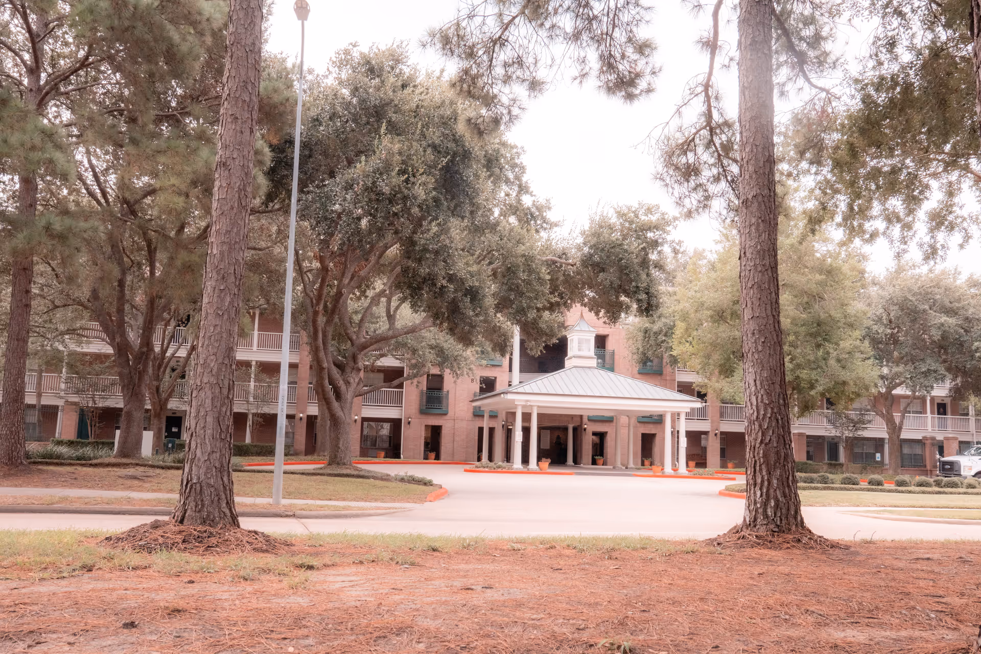 Front exterior view of a multi-story brick building with a covered entrance supported by white columns, surrounded by large trees and a driveway with red curbs.