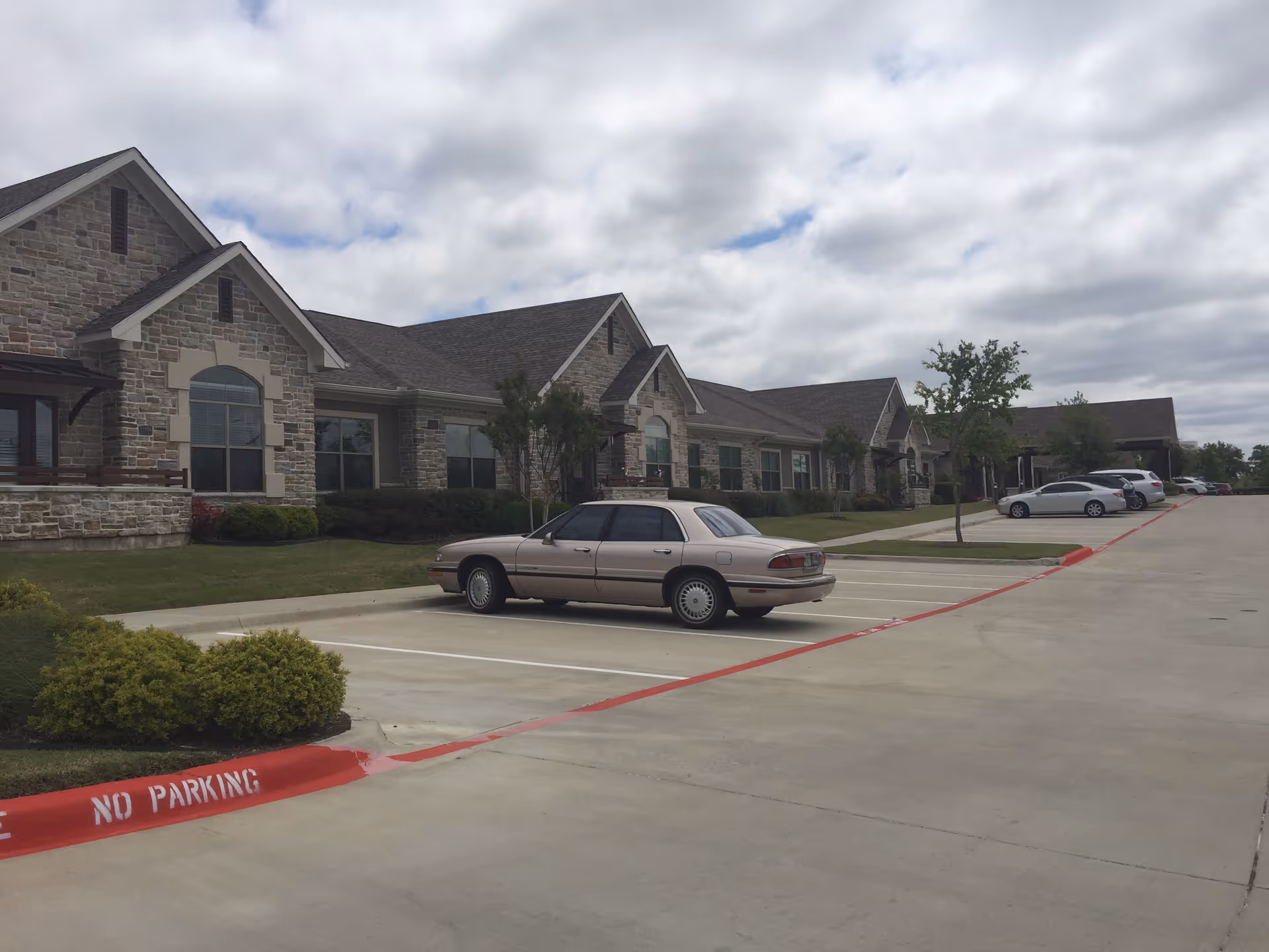 A stone facade senior living building along a curved driveway with parked cars.