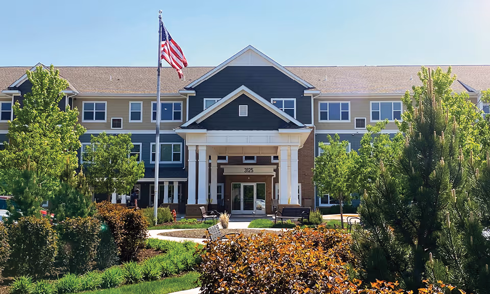 Two-story senior living building with central entrance, American flag, and landscaped front yard.