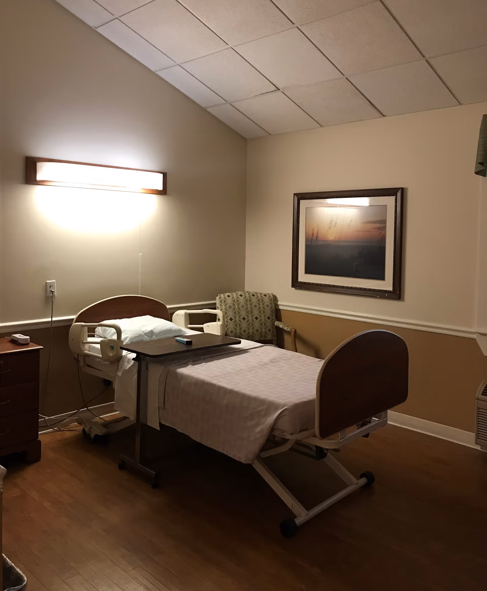 A hospital-style bed with white sheets and a pillow in a room with beige walls and wooden flooring. There is a wooden nightstand with a remote control on it to the left of the bed. A green patterned armchair is positioned against the wall near the bed. Above the chair, there is a framed picture of a sunset or sunrise. A wall-mounted light fixture illuminates the room from above the bed.