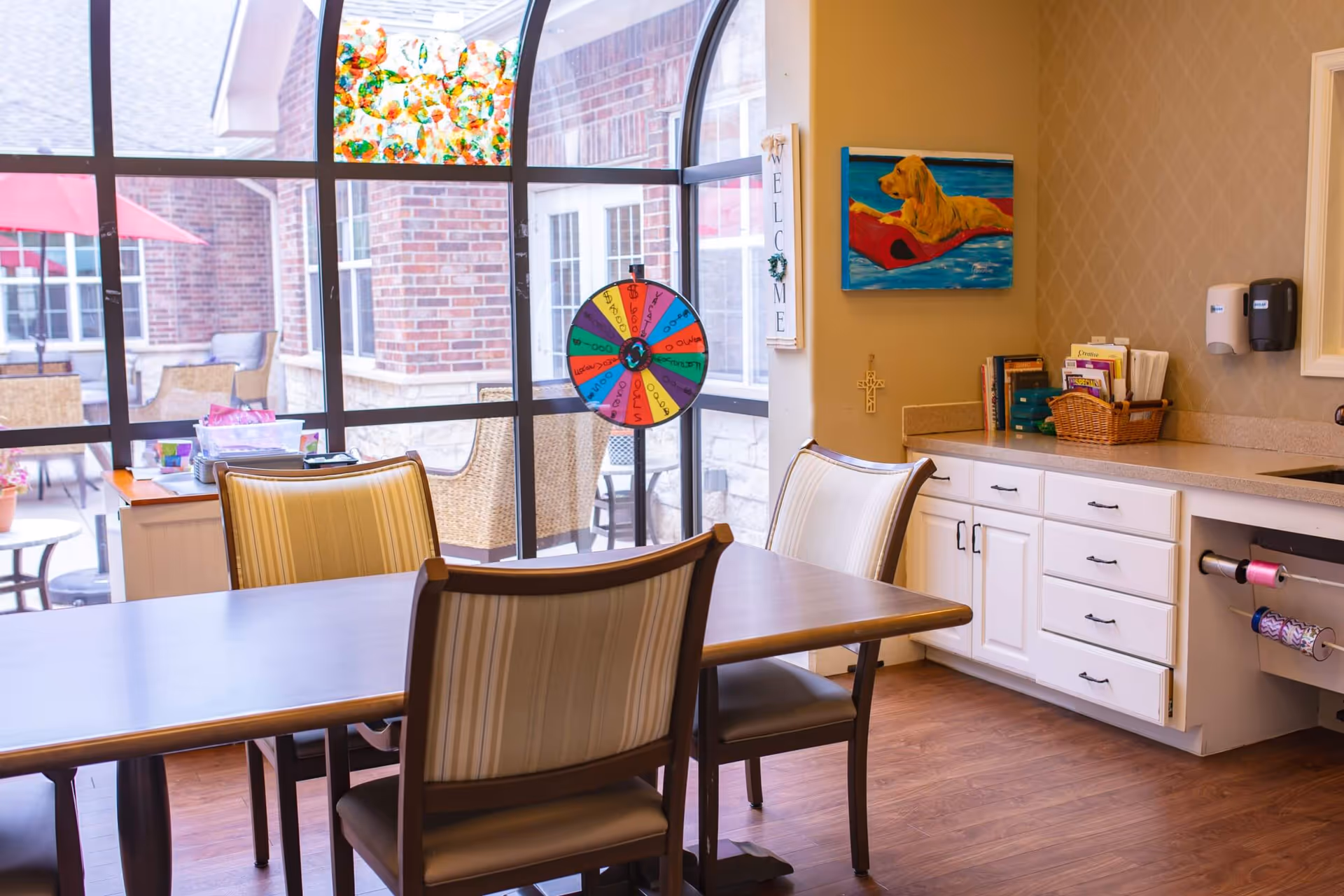A bright room with a wooden table and four striped cushioned chairs. Behind the table is a large window with a view of an outdoor patio area with wicker chairs and a red umbrella. Inside the room, there is a colorful spinning wheel, a welcome sign on the wall, a painting of a dog on a red float in water, and white cabinets with various items on the countertop including books and a basket.