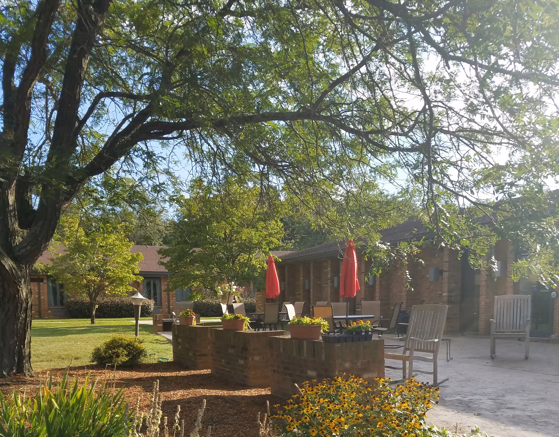 Outdoor courtyard with brick seating walls, tables, chairs, and red umbrellas among trees.