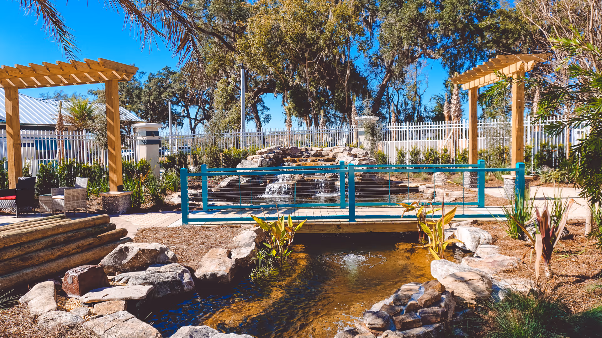 Outdoor courtyard with a small pond and rock waterfall, a turquoise footbridge, pergolas, and seating areas.