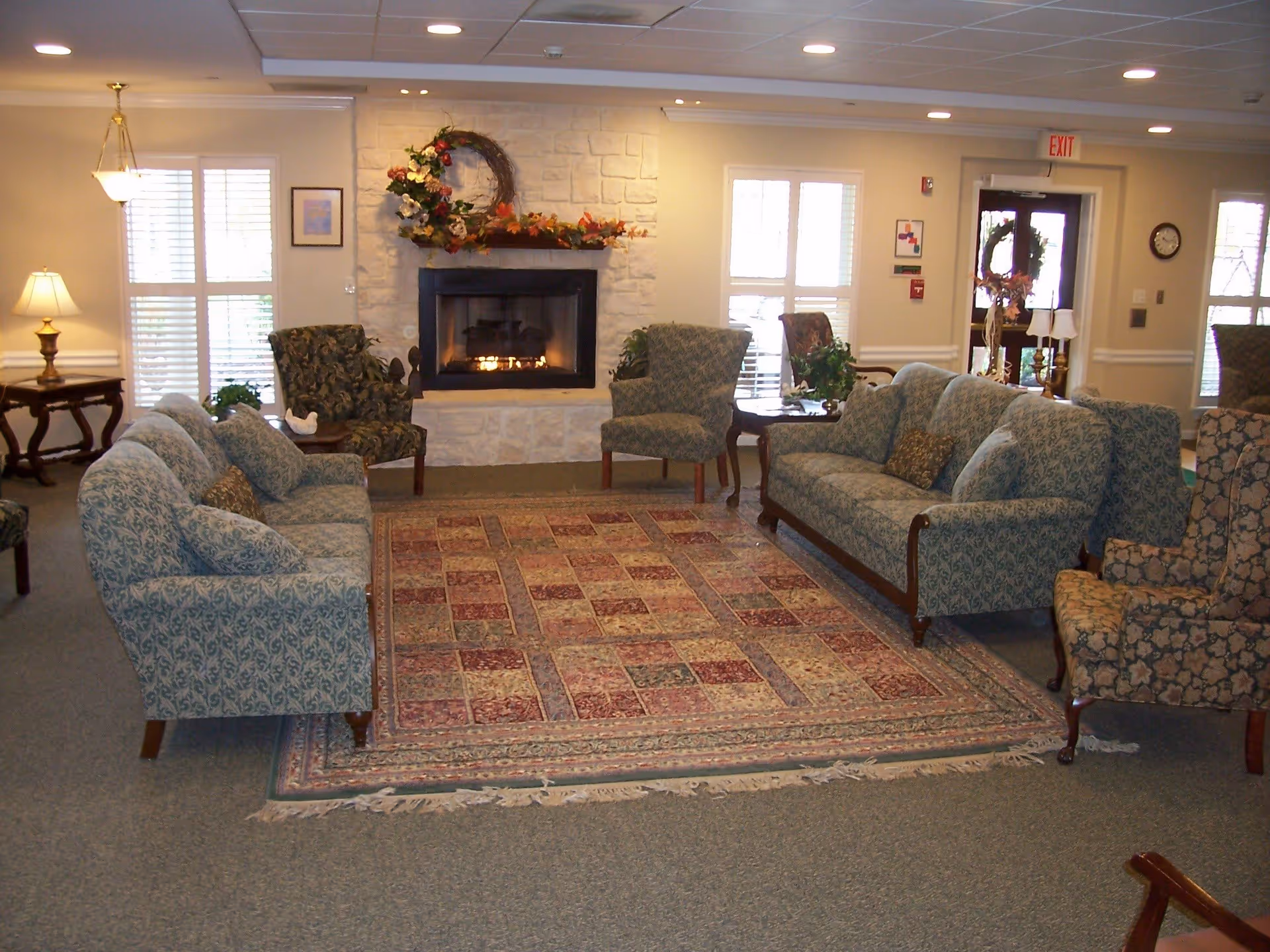 A cozy senior living facility common area with two patterned sofas and several armchairs arranged around a large decorative rug. A stone fireplace with a floral wreath and autumn decorations is centered on the far wall. The room is softly lit with table lamps and natural light coming through windows with white shutters. There is an exit door with a wreath and a clock on the wall.