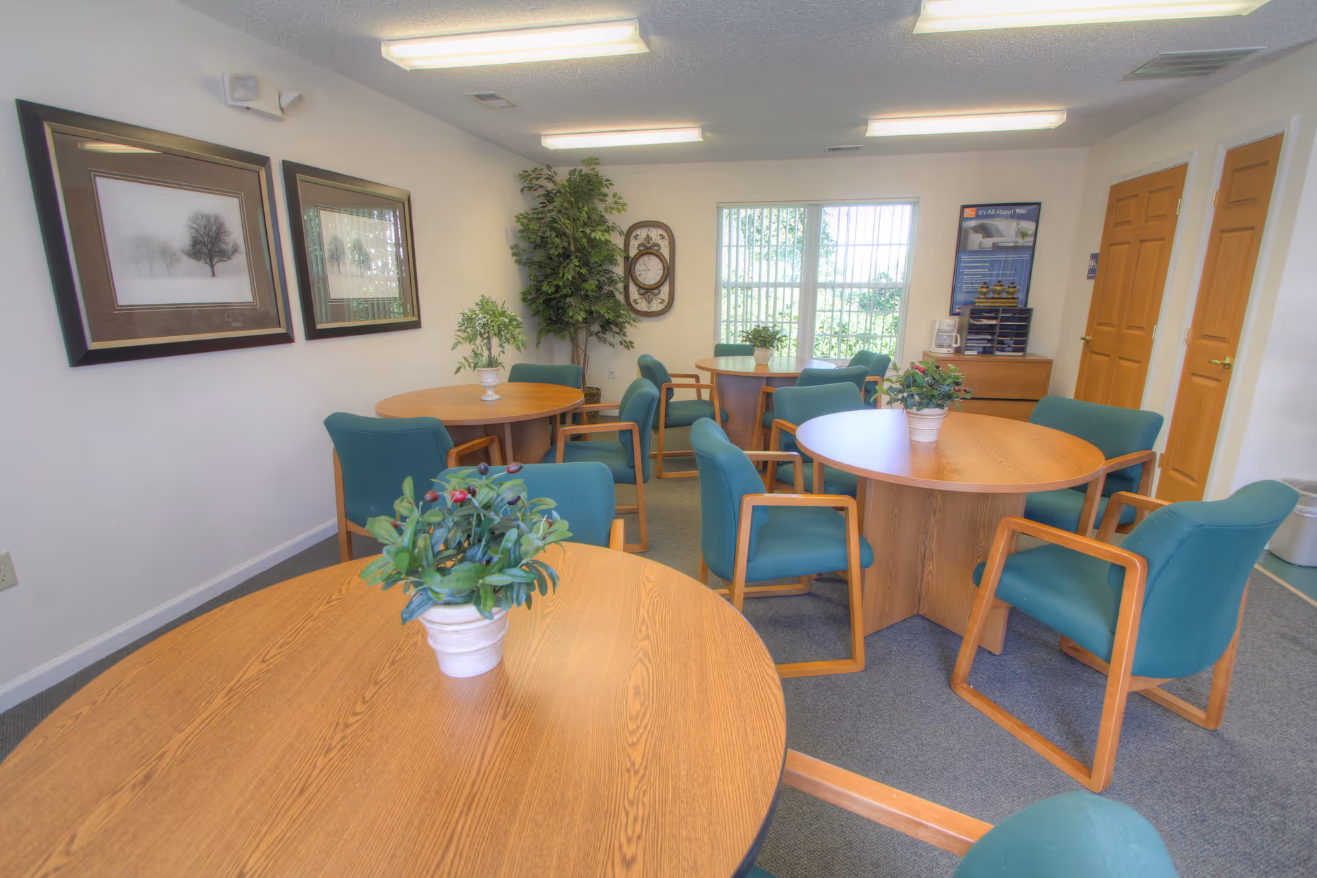 Round wooden tables with teal chairs fill a bright senior living facility common area.
