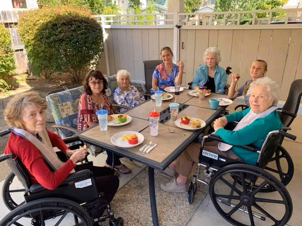A group of elderly women and a caregiver sitting around an outdoor table enjoying a meal together. The setting is a fenced patio area with greenery in the background. Some of the women are in wheelchairs, and there are plates of food and colorful cups on the table.
