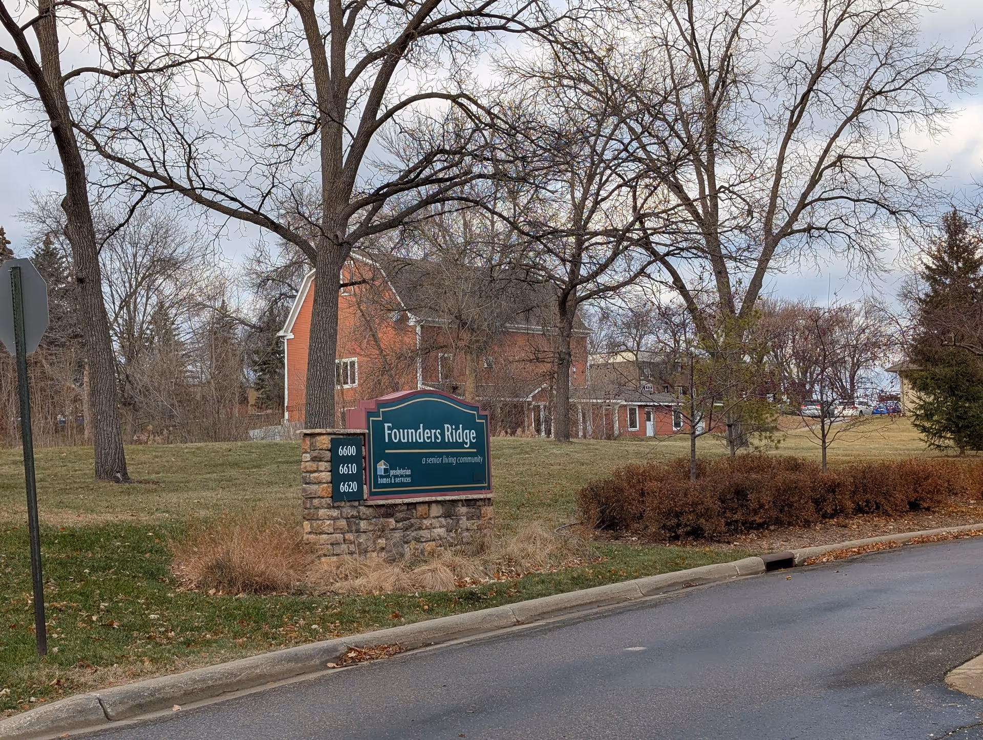 Stone entrance sign reading "Founders Ridge" on a grassy lawn with leafless trees and a red brick building in the background.